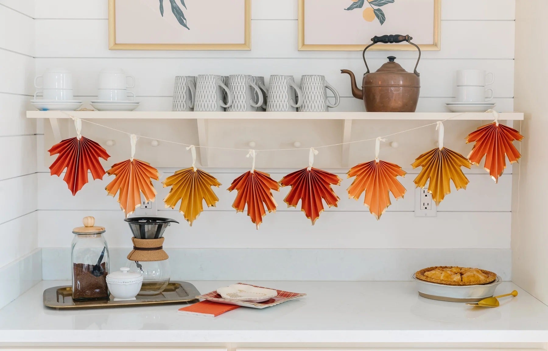 A kitchen counter decorated with maple leaf paper party banner from Momo Party. Underneath are some coffee brewing set and homemade baked pie, a very autumn fall home decor idea for the harvest season.