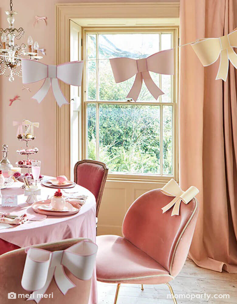 An elegant and chic bow themed party set up featuring Momo Party's pastel heart shaped plates and napkins and 3D bow party cups on the table, above the table there's an elegant chandelier with Meri Meri's 3D bow garland hung over. On the table there are scrumptious treats and sweets for tea time. The chairs are also adorn with 3D bow decoration for a playful touch.
