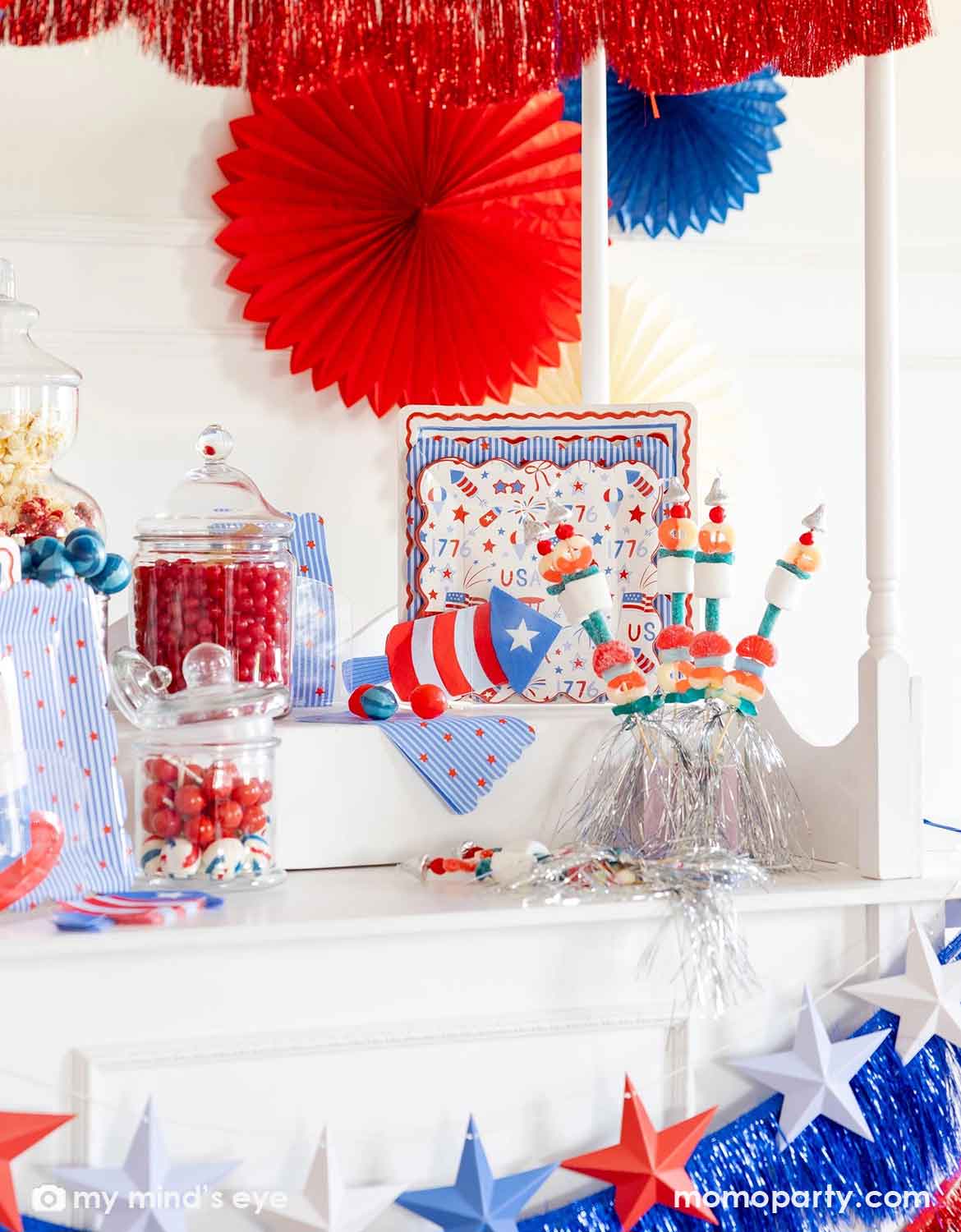 A close up shot of a patriotic themed decorated dessert cart with various American themed decors and tablewares in red, white, and blue from Momo Party, including paper fans, fringe bunting garlands, star party banners, USA themed plates, napkins and cups. Along with candy jars and decorative marshmallows in firecracker shapes, making this a perfect inspiration for a festive table setting for a kid's friendly Fourth of July party, Memorial Day celebration or a summer gathering.