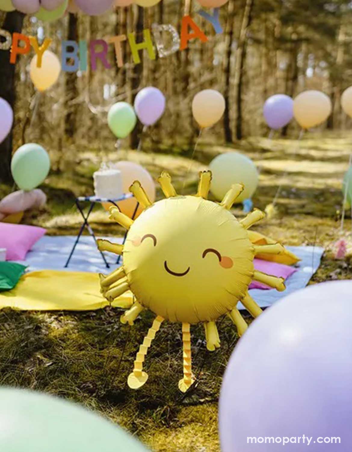 Happy birthday decoration with Momo Party's 41" sun-shaped foil balloon, floating pastel colored latex balloons in the back and a multicolor happy birthday party banner hung above at an outdoor party on a lawn in woods.