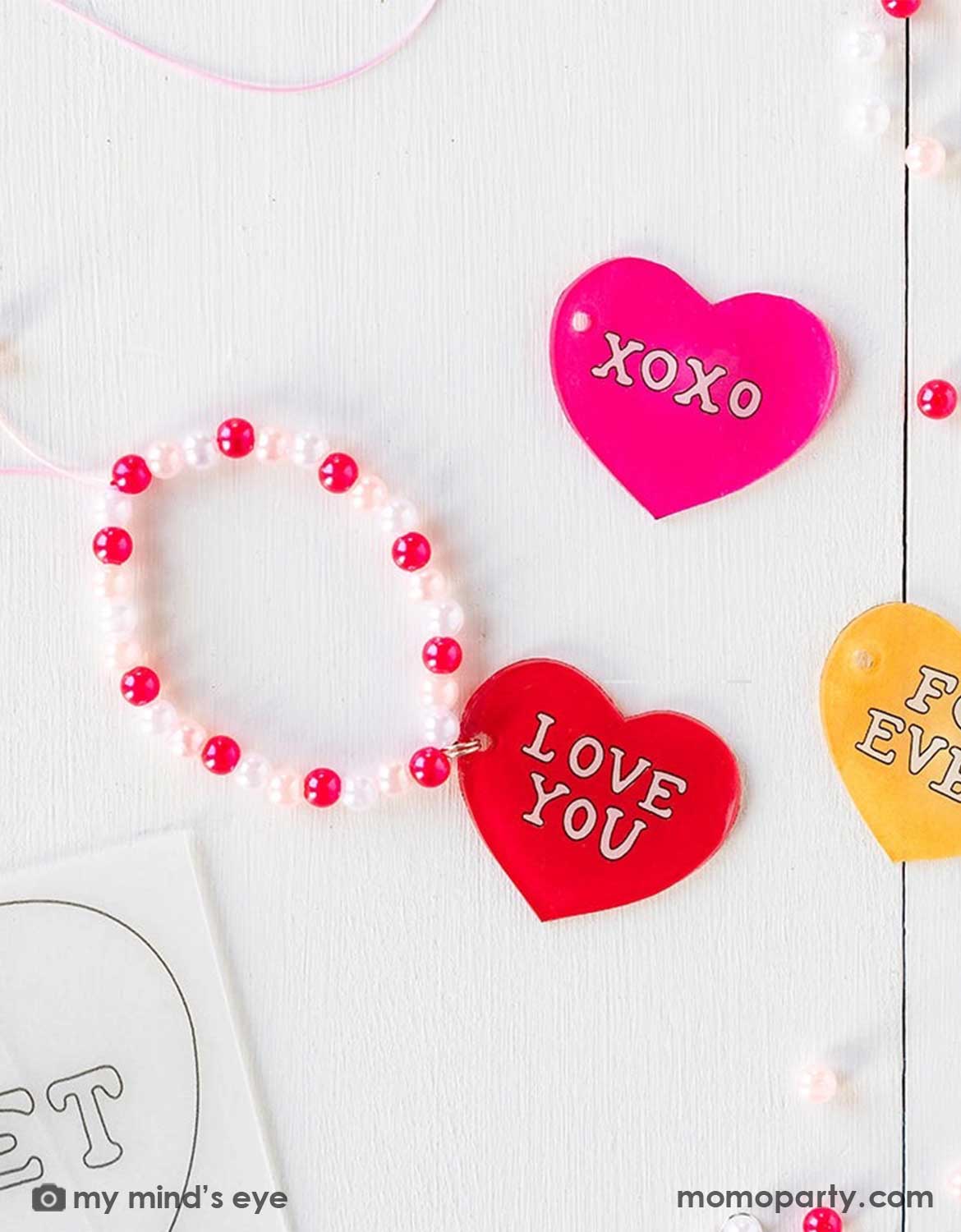 Pink and red heart-shaped bracelet charm with 'LOVE YOU' and 'XOXO' text and beads on a light background.