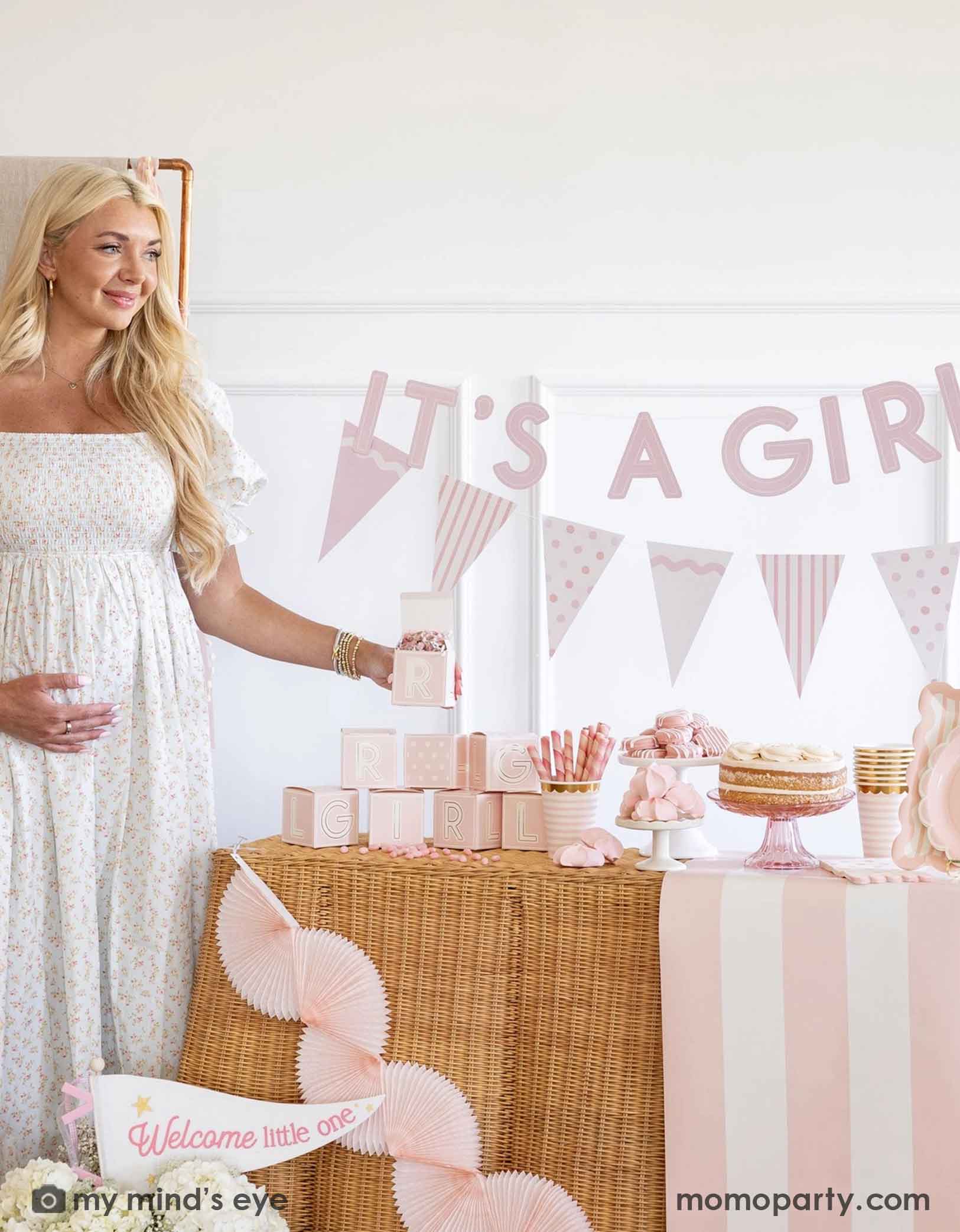 A pregnant woman standing next to a baby shower table with 'It's a Girl' banner and baby pink shower decorations and party supplies from Momo Party including tablewares, party banners, table runner and bunting garlands.