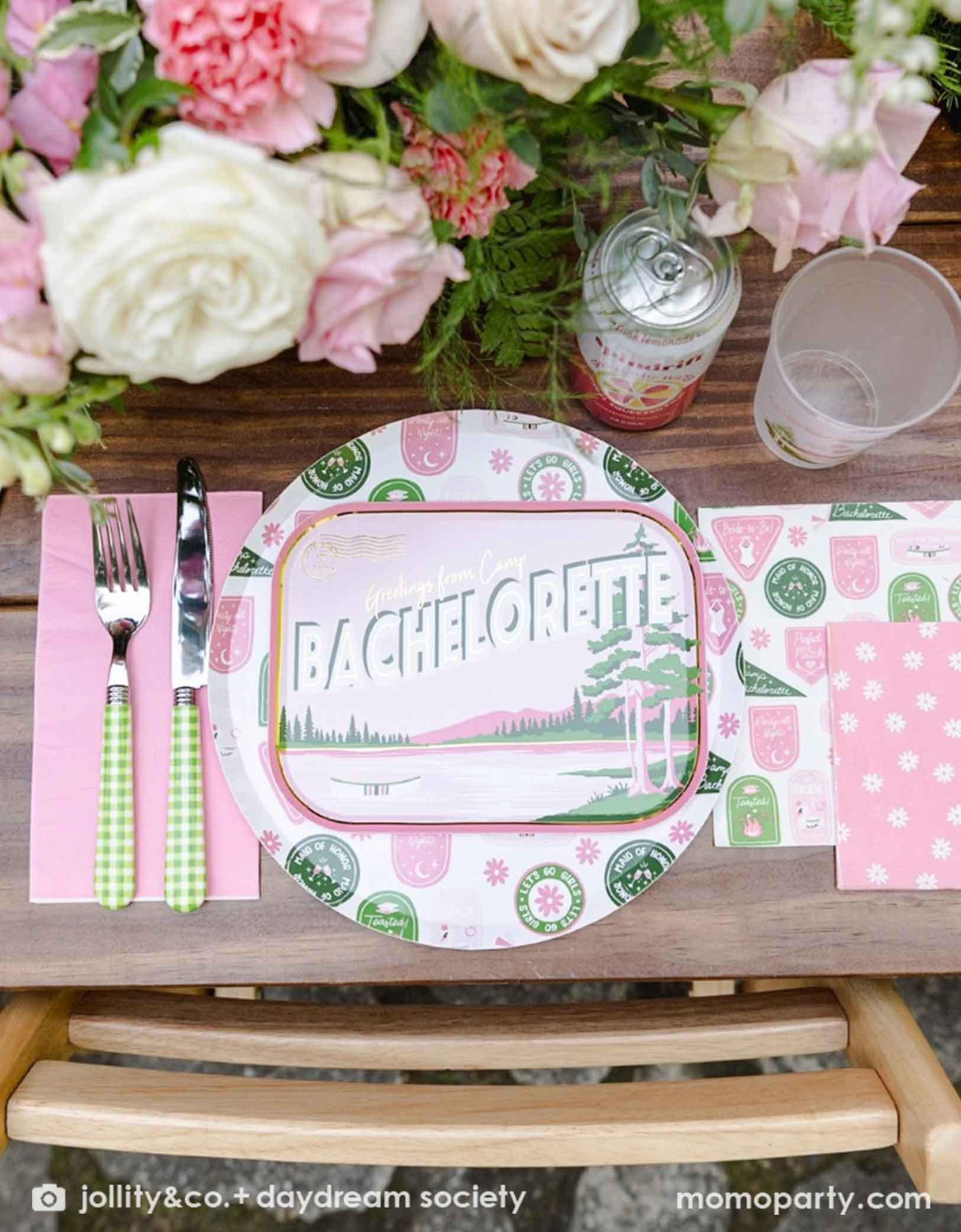 A beautiful bachelorette party table setting with a Camp Bachelorette themed plate, napkins and cutlery from Momo Party, along with flowers, drinks and cups on a wooden table.