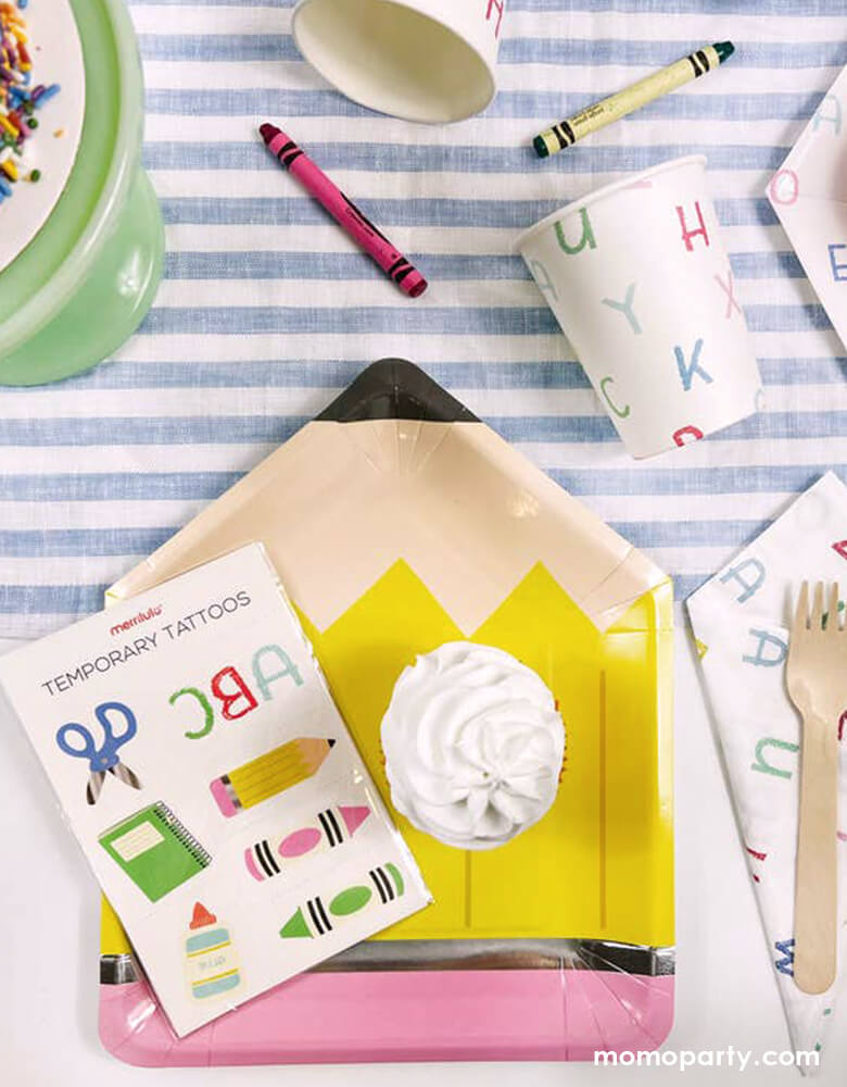 A back to school party table filled with Momo Party's school themed party supplies including a pencil shaped plate, ABC letter patterned napkins and party cups, on top of the plate is a temporary tattoos sheet featuring colorful school themed illustrations. All this makes a great party inspo for kid's back to school party or first day of school celebration.