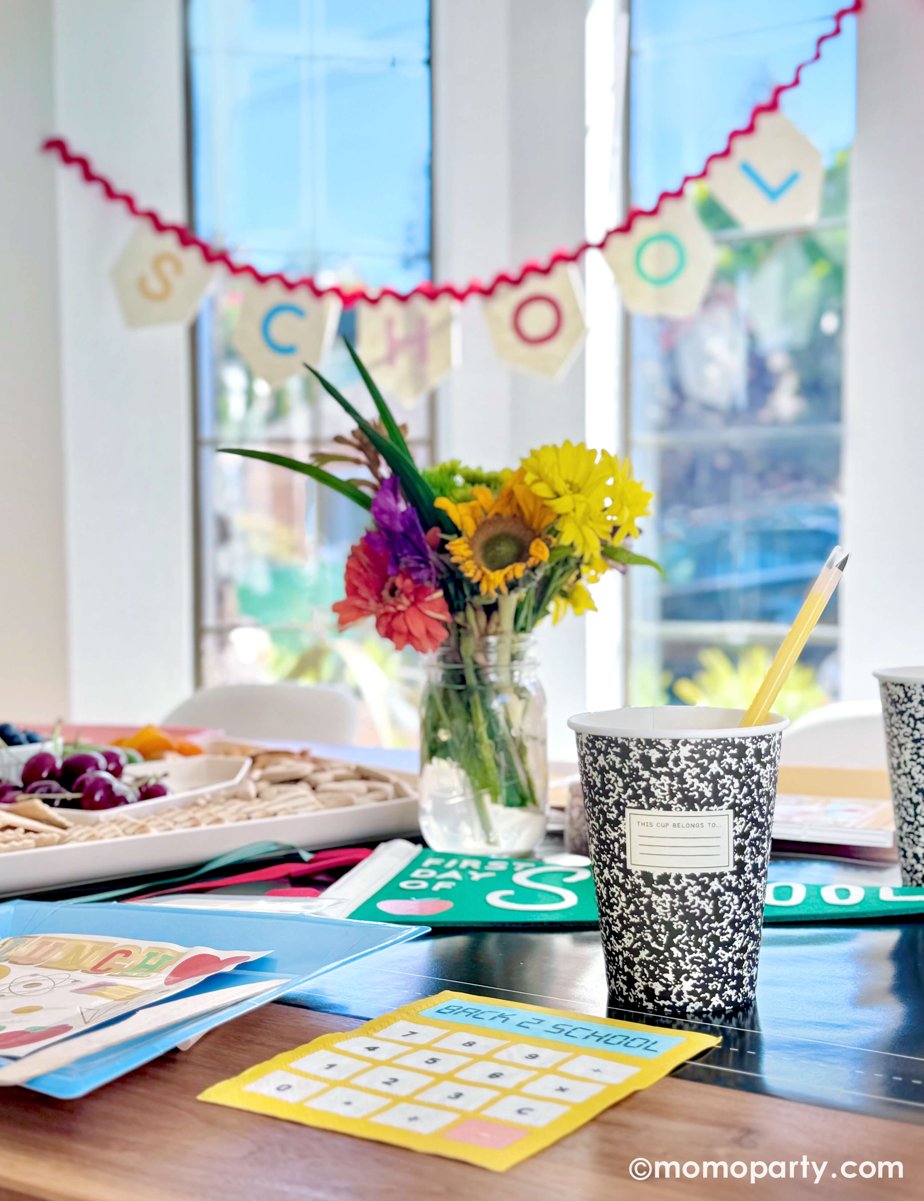 A kids' back to school party table setting featuring various school themed party decors and tableware from Momo Party including notebook party cups with pencil reusable straws, calculator shaped dinner napkins, lunchbox shaped plates and apple first day of school party pennant, with a summer flower arrangement and a kid's snack board, along with "School Day" party garland, this simple yet festive set up is perfect to celebrate your child's first day of school!