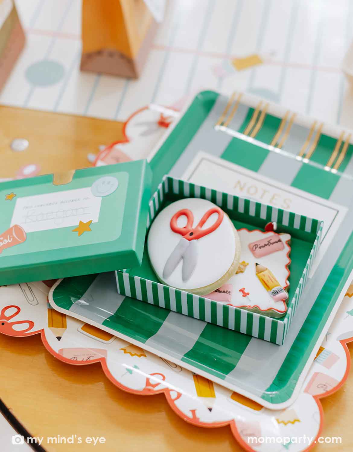 A styled flat-lay of a first day of school back to school celebration featuring a green and white striped school themed treat box containing a round sugar cookie decorated with a white icing background and red-orange scissors, alongside matching school themed papery plates on a school lined table runner on a wooden table surface.