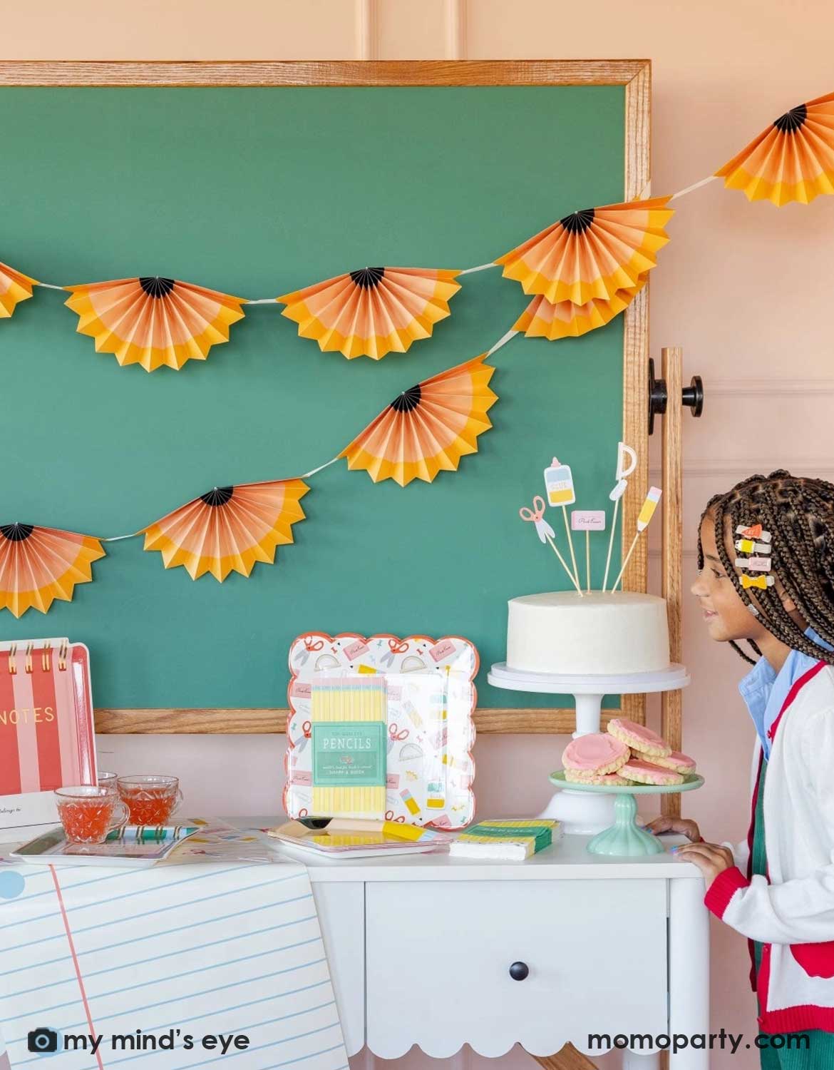 A girl in her uniform and school themed hair clips standing next to a school themed decorated party table with a cake, surrounded by various school themed party supplies and decorations from Momo Party including party plates, napkins, table runner in front of a chalkboard above which is decorated with pencil party fans garland hung above, making this perfect inspiration for kid's first day of school celebration or back to school party.