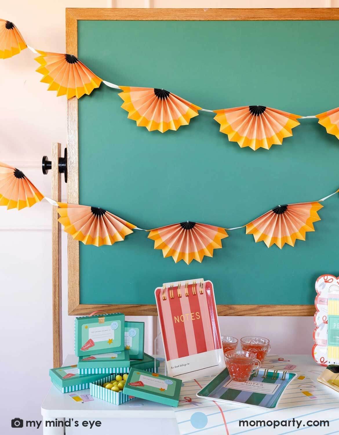 A school themed party in a classroom setting featuring a table with various school themed party supplies and decorations from Momo Party including notebook shaped party plates, napkins, lunchbox treat boxes on a table runner in front of a chalkboard which is decorated with pencil party fan garland hung above, making this perfect inspiration for kid's first day of school celebration or back to school party.