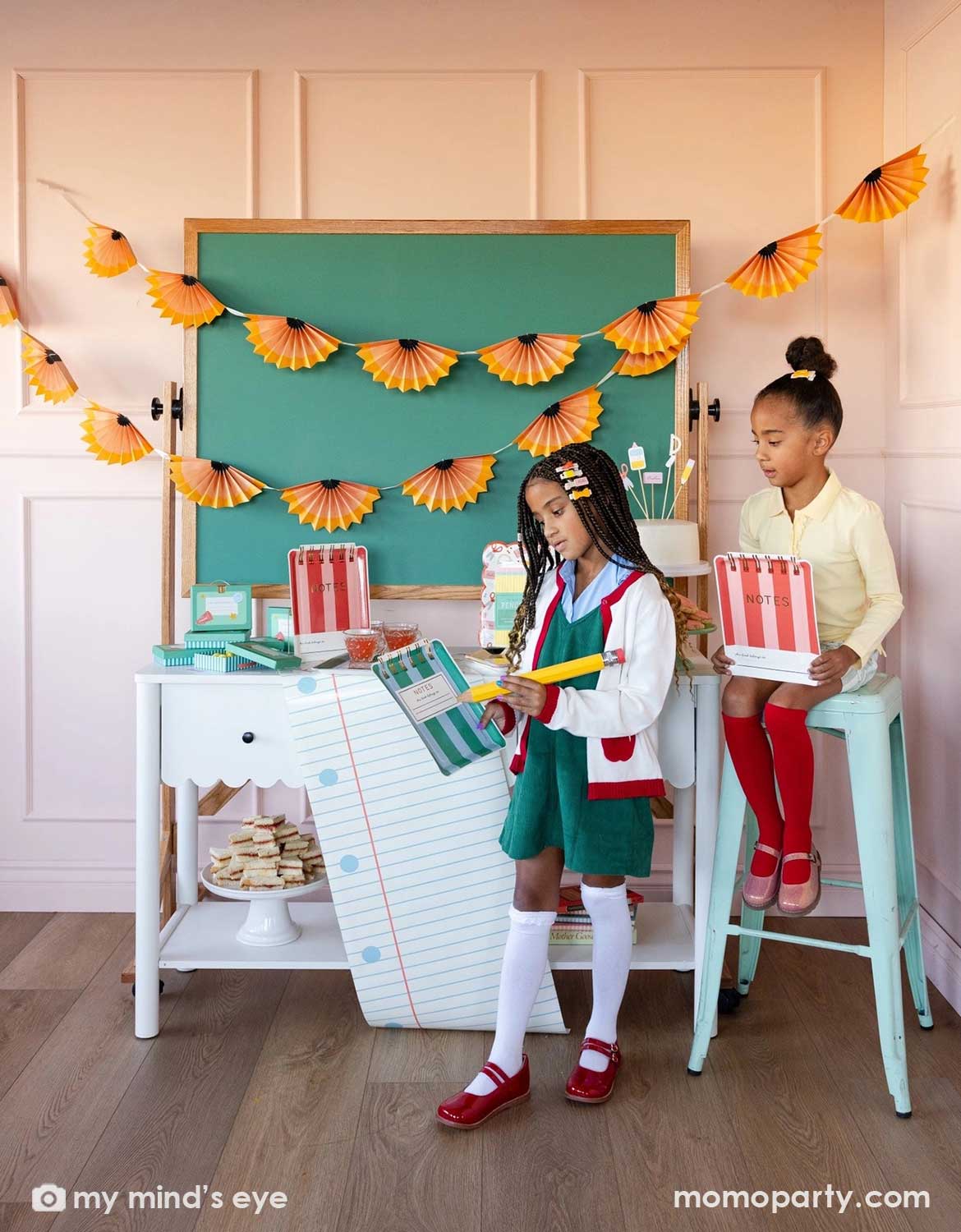 Two girls holding notebook shaped party plates and a giant pencil sitting and standing in front of a school themed decorated party table with a cake, surrounded by various school themed party supplies and decorations from Momo Party including party plates, napkins, table runner in front of a chalkboard above which is decorated with pencil party fans garland hung above, making this perfect inspiration for kid's first day of school celebration or back to school party.