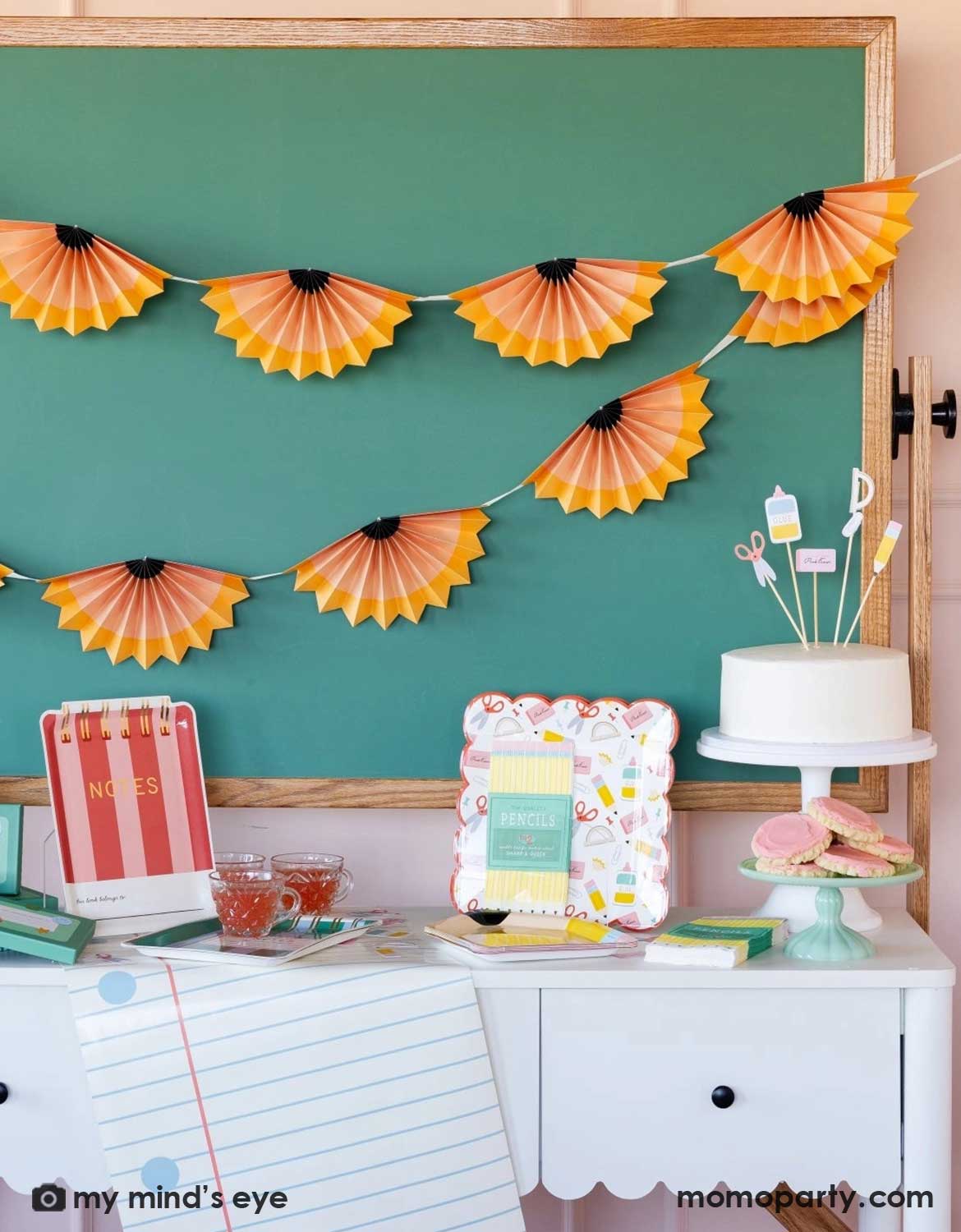 A school themed party in a classroom setting featuring a table with various school themed party supplies and decorations from Momo Party including school supplies icon party plates, red notebook shaped plates, napkins on a table runner. Next to it is a cake topped with school supplies toppers along with some cookies in front of a chalkboard which is decorated with pencil party fan garland hung above, making this perfect inspiration for kid's first day of school celebration or back to school party.