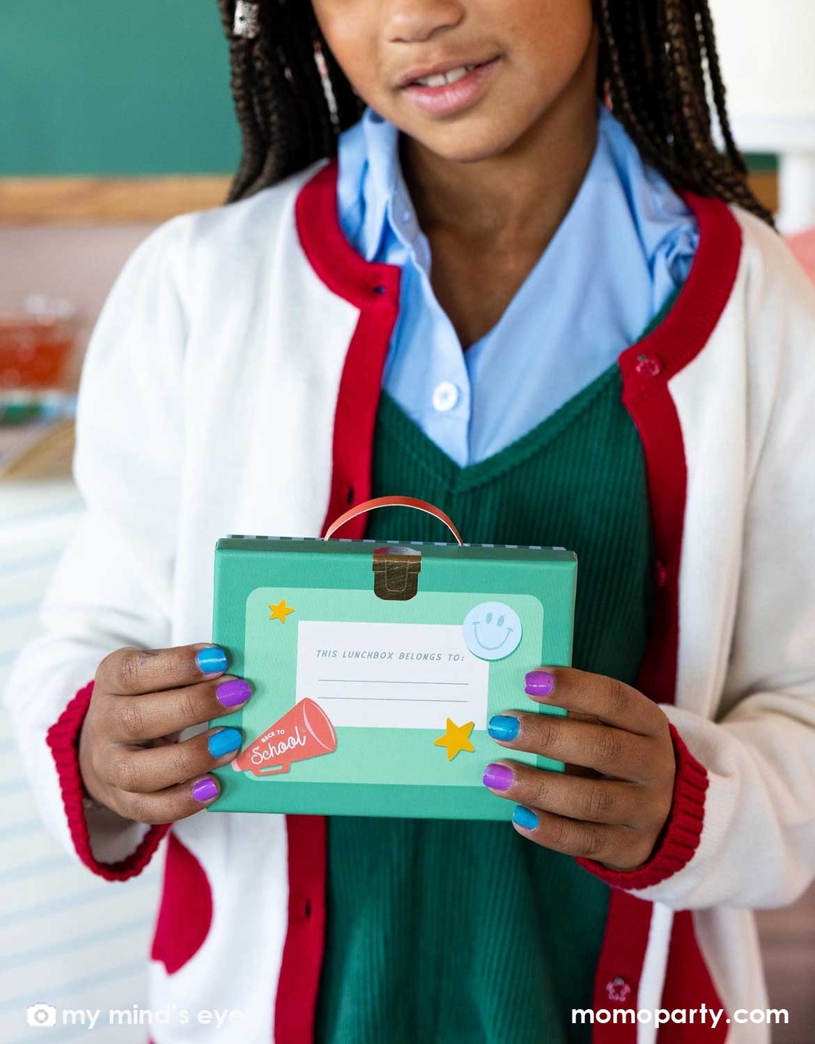 A girl in school uniform holding Momo Party's lunch box shaped treat box standing in a classroom setting.