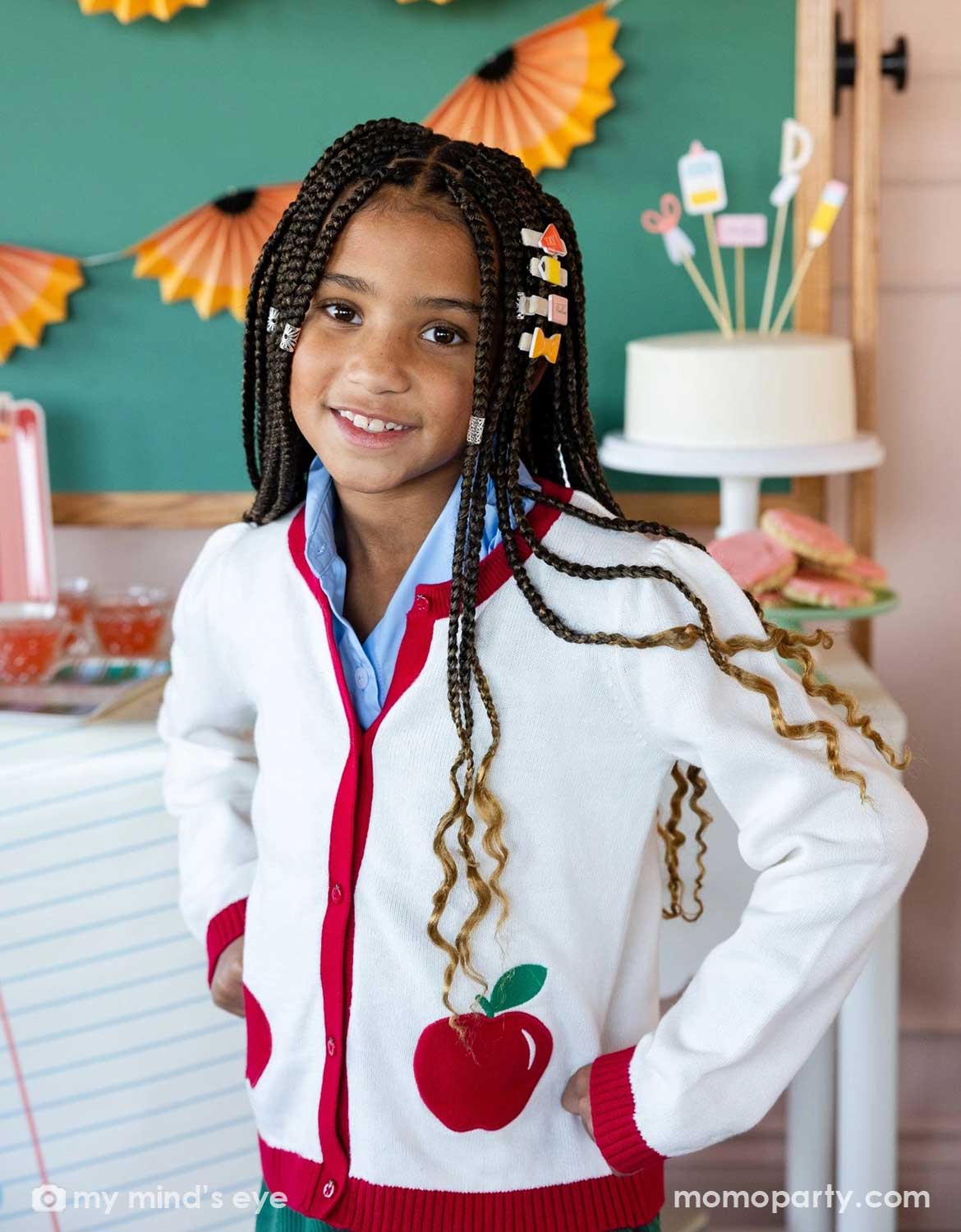 A young girl in a white cardigan with an apple design and her uniform with braids adorned with Momo Party's colorful back to school hair clips with school supplies themed design, standing in a decorated room.