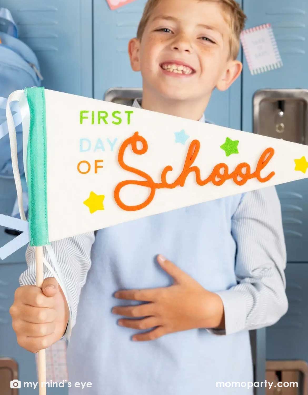 A boy holding Momo Party's 'First Day of School' felt party pennant in front of blue lockers in a classroom setting.