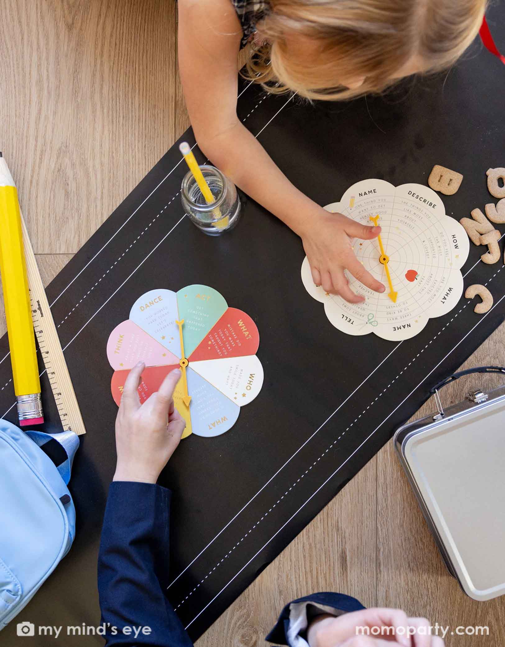 Two school-aged kids playing an activity spinning game on the floor covered with Momo Party's chalkboard paper table runner. On the runner there are jumbo school supplies including a ruler, a jumbo pencil, some alphabet cookies, a lunch box and its bag.