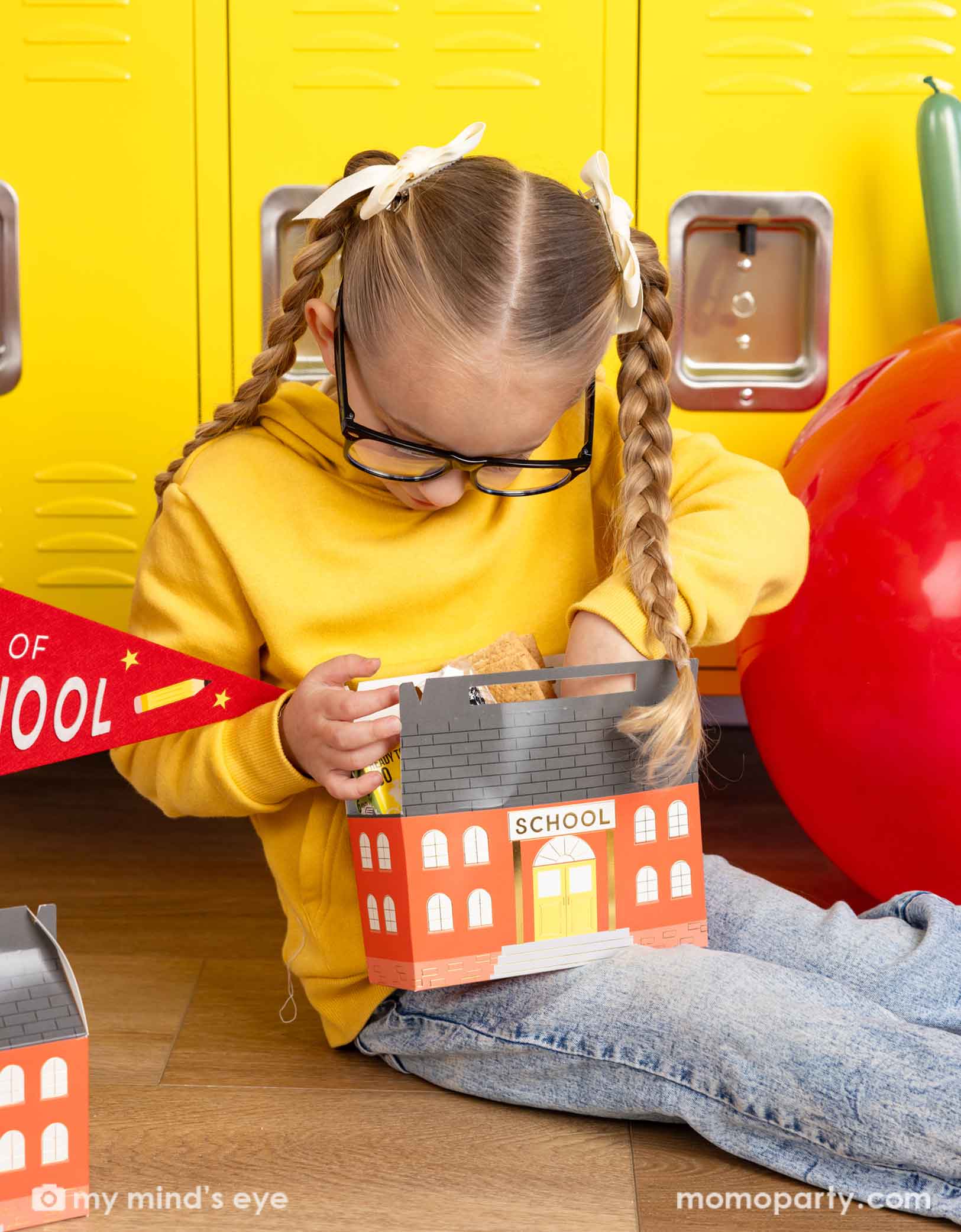 A girl in her yellow sweatshirt and glasses looking into Momo Party's school building shaped treat box. She is sitting in front of a first day of school celebration decoration featuring various school themed party decors from Momo Party including a jumbo apple latex balloon, first day of preschool party pennant, and school shaped treat boxes.