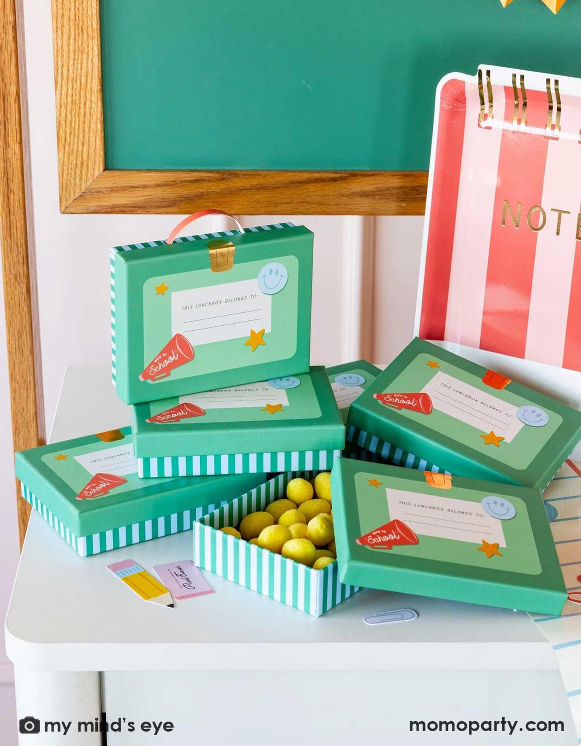 A bunch of Momo Party's lunch box shaped treat boxes filled with colorful candies and lollipops stacked up on a school themed party table featuring various of school themed party supplies from Momo Party including red striped notebook shaped plate, school supplies confetti and notebook blue line paper table runner, making this a perfect tablescape inspiration for kid's school themed party or first day of school celebration.