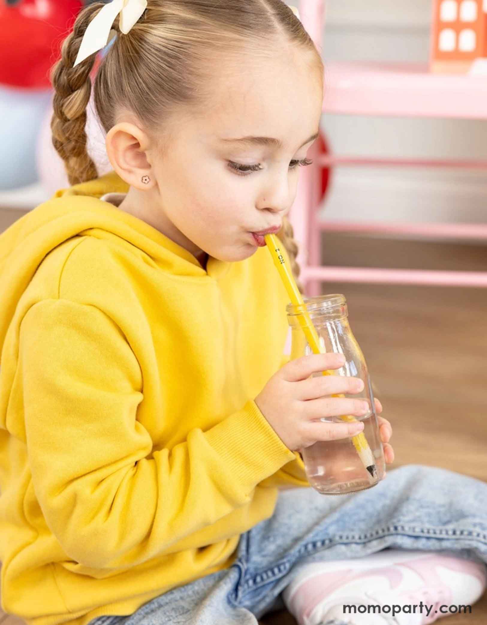A little girl wearing a yellow sweatshirt sipping water with Momo Party's pencil shaped straw from a milk bottle. This eco-friendly reusable straw is perfect for your party and come in a fun pencil shape. It's perfect for kid's back to school party or first day of school celebration.