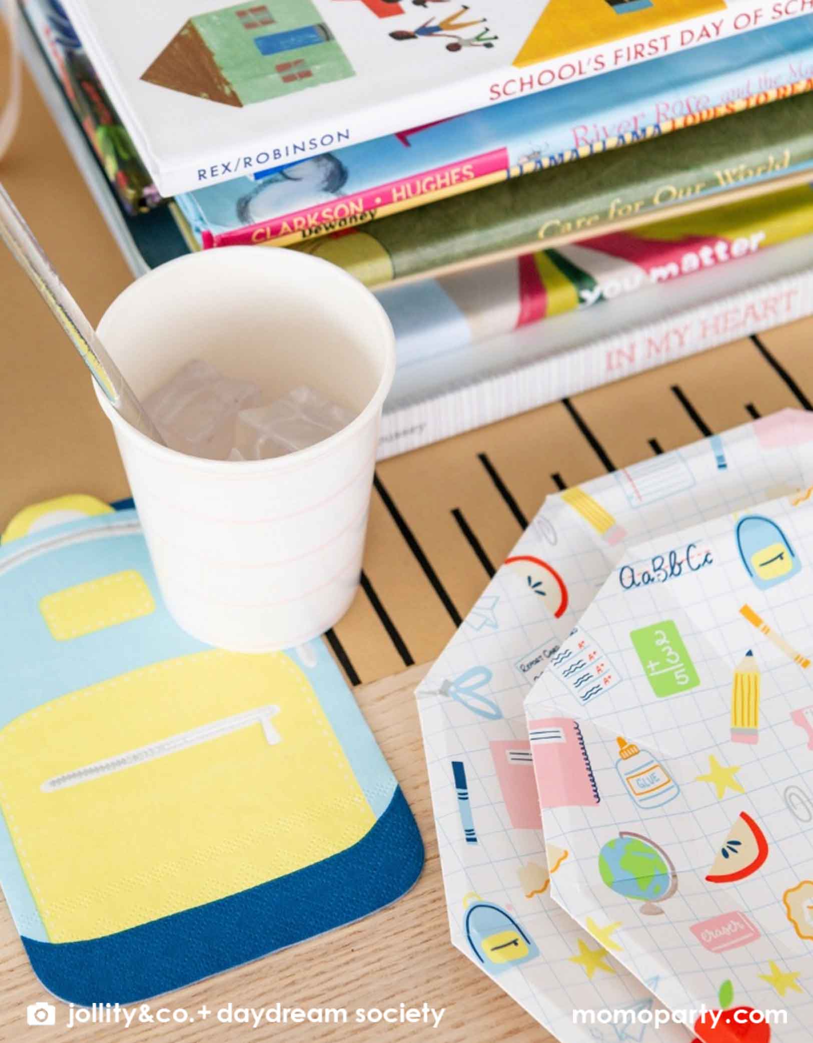 A close up shot of an adorable kid's school themed party table featuring school supplies themed party tablewares from Momo Party including the Smarty Pants backpack shaped napkin, a pink striped party cup, along with large and small school icons patterned plates, all on a ruler inspired table runner. In the middle of the table there is a pile of books as centerpiece, making this a great decoration inspiration for kid's first day of school celebration or a back to school party.