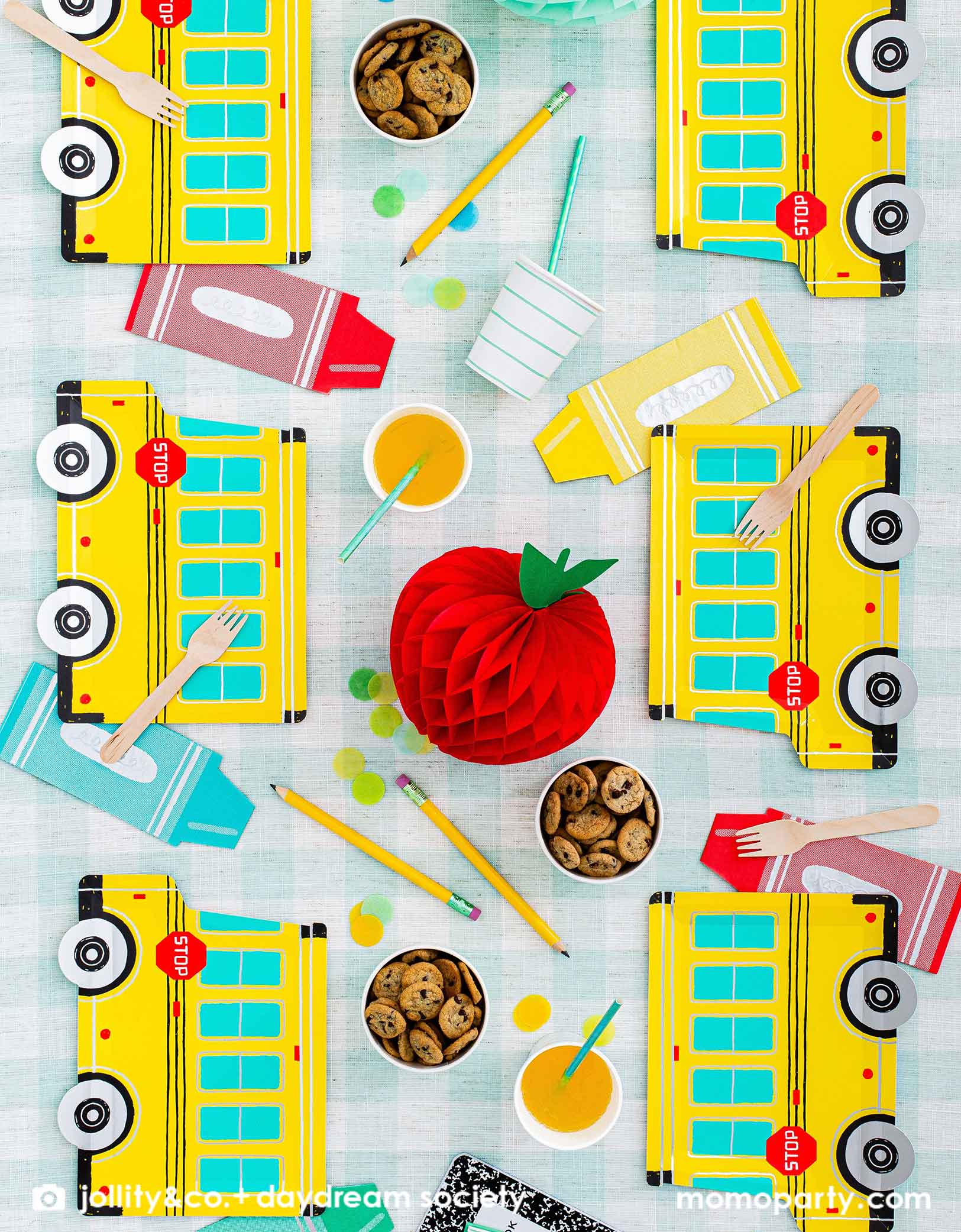 An overhead shot of a festive back to school party table featuring Momo Party's colorful school themed tableware including Daydream Society's school bus shaped plates, crayon shaped napkins with aqua striped party cups and snack cups filled with cookies, all on a mint gingham checkered tablecloth. With an apple shaped honeycomb decoration, festive confetti and pencils on the party table, this gives a great idea and inspiration for a kid's back to school party or a first day of school celebration.