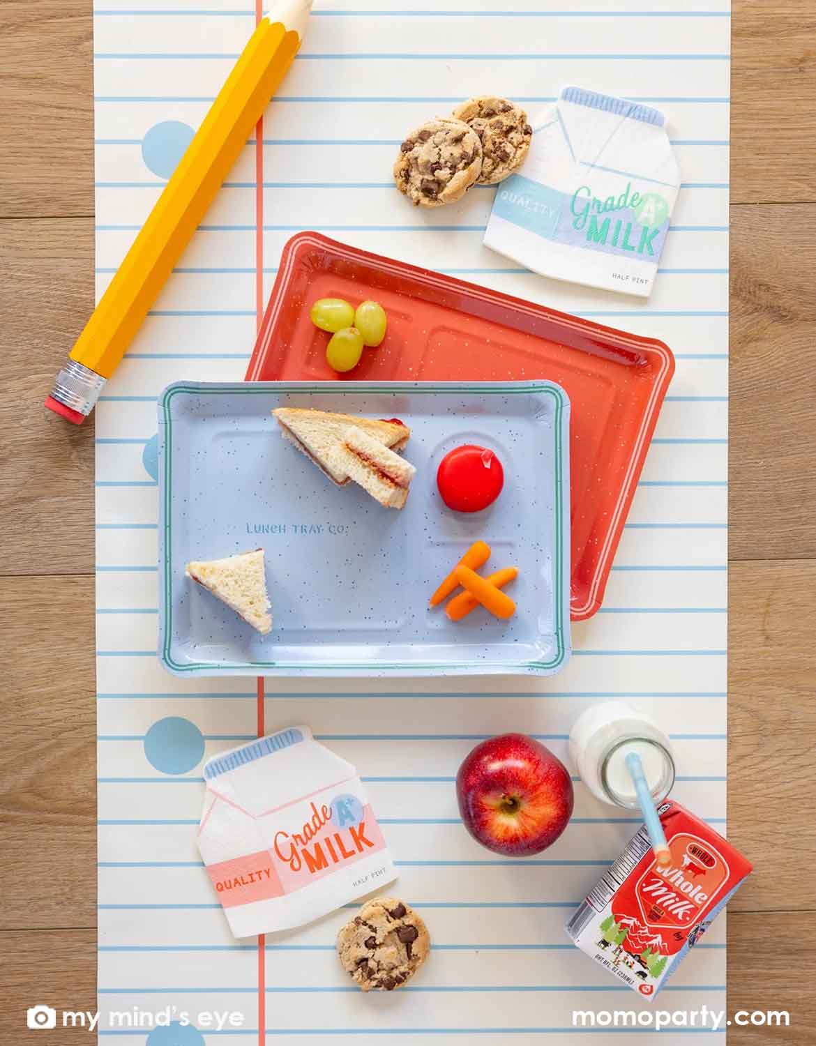 An overhead shot of a school themed party table featuring Momo Party's colorful lunch box shaped plates, milk bottle shaped napkins, a large pencil and lunch food and drinks spread across the notebook blue line paper table runner, making this a perfect back to school party or first day of school celebration tablescape inspiration.