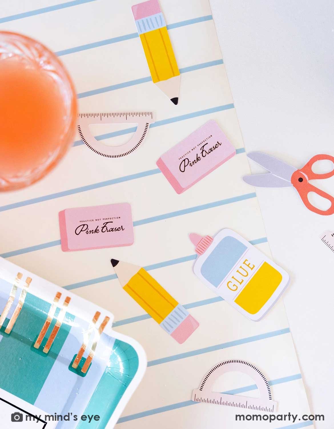 A close up shot of Momo Party's school supplies themed party confetti with pink erasers, glue, and scissors by My Mind's Eye scattered across on a blue striped table runner.