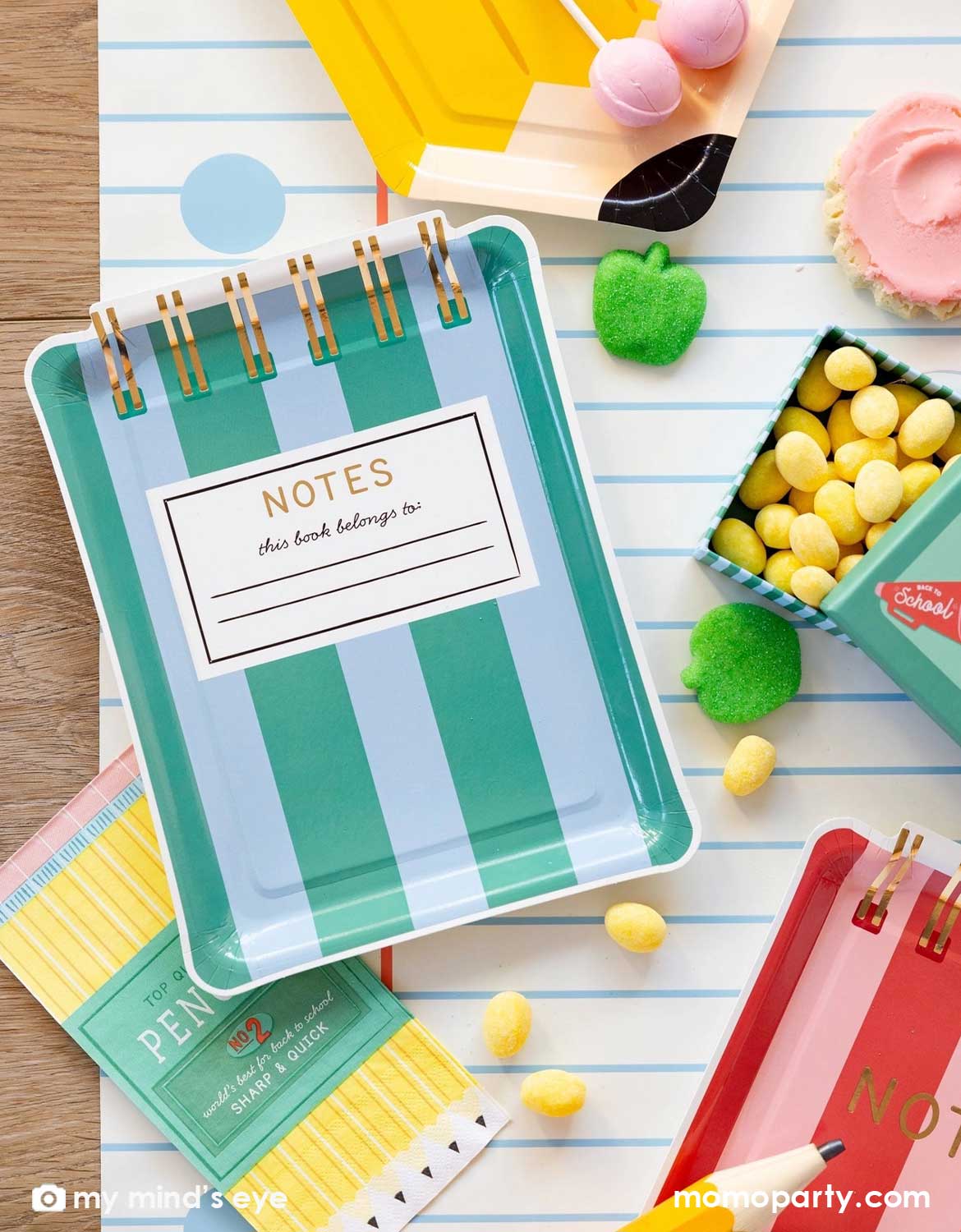An overhead shot of a school themed party table featuring Momo Party's green striped notebook shaped plate, pencil set napkins and small lunch box shaped treat boxes filled with candies, pencil shaped plate with green apple shaped gummies and cupcakes spread across the notebook blue line paper table runner, making this a perfect back to school party or first day of school celebration tablescape inspiration.