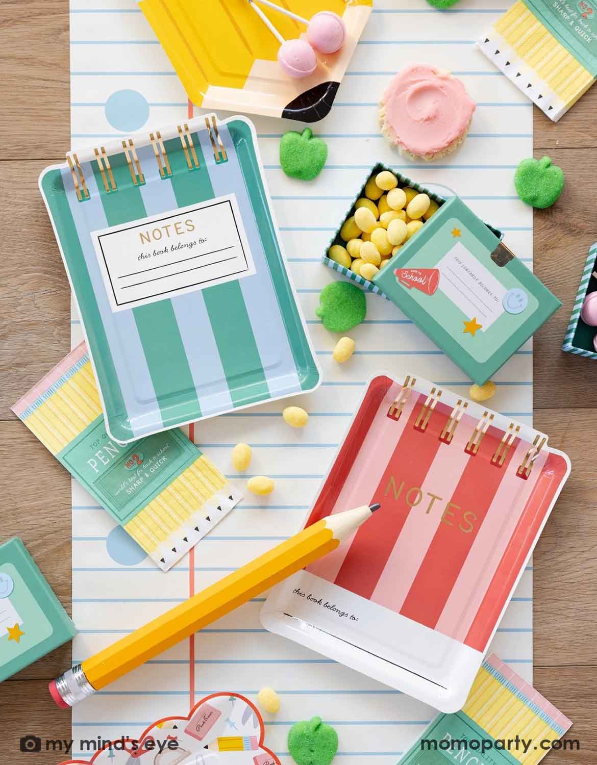 An overhead shot of a school themed party table featuring Momo Party's colorful striped notebook shaped plates, pencil set napkins, pencil shaped plates and small lunch box shaped treat boxes filled with candies, with green apple shaped gummies and cupcakes spread across the notebook blue line paper table runner, making this a perfect back to school party or first day of school celebration tablescape inspiration.