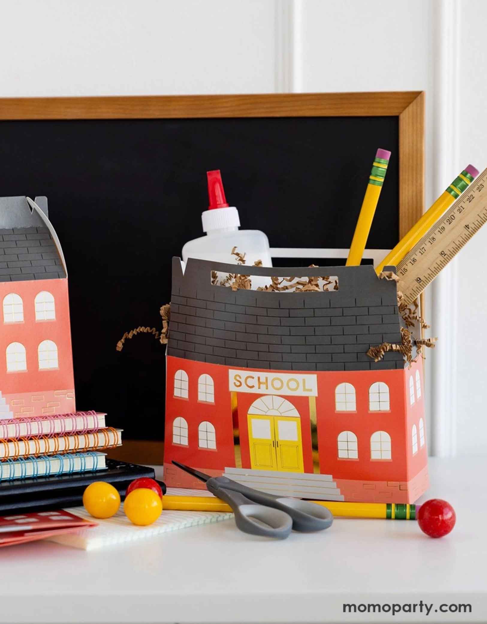 Two of Momo Party's 7" x 2.5" x 5.5" School House Treat Boxes by My Mind's Eye on a desk with school supplies including pencils, rulers, scissors, glue bottles in them. Around the treat boxes there are some colorful notebooks and some red and yellow gum balls and in the back there's a small chalkboard, making this an adorable set up for kid's school themed celebration, be it a back to school party or a first day of school celebration.