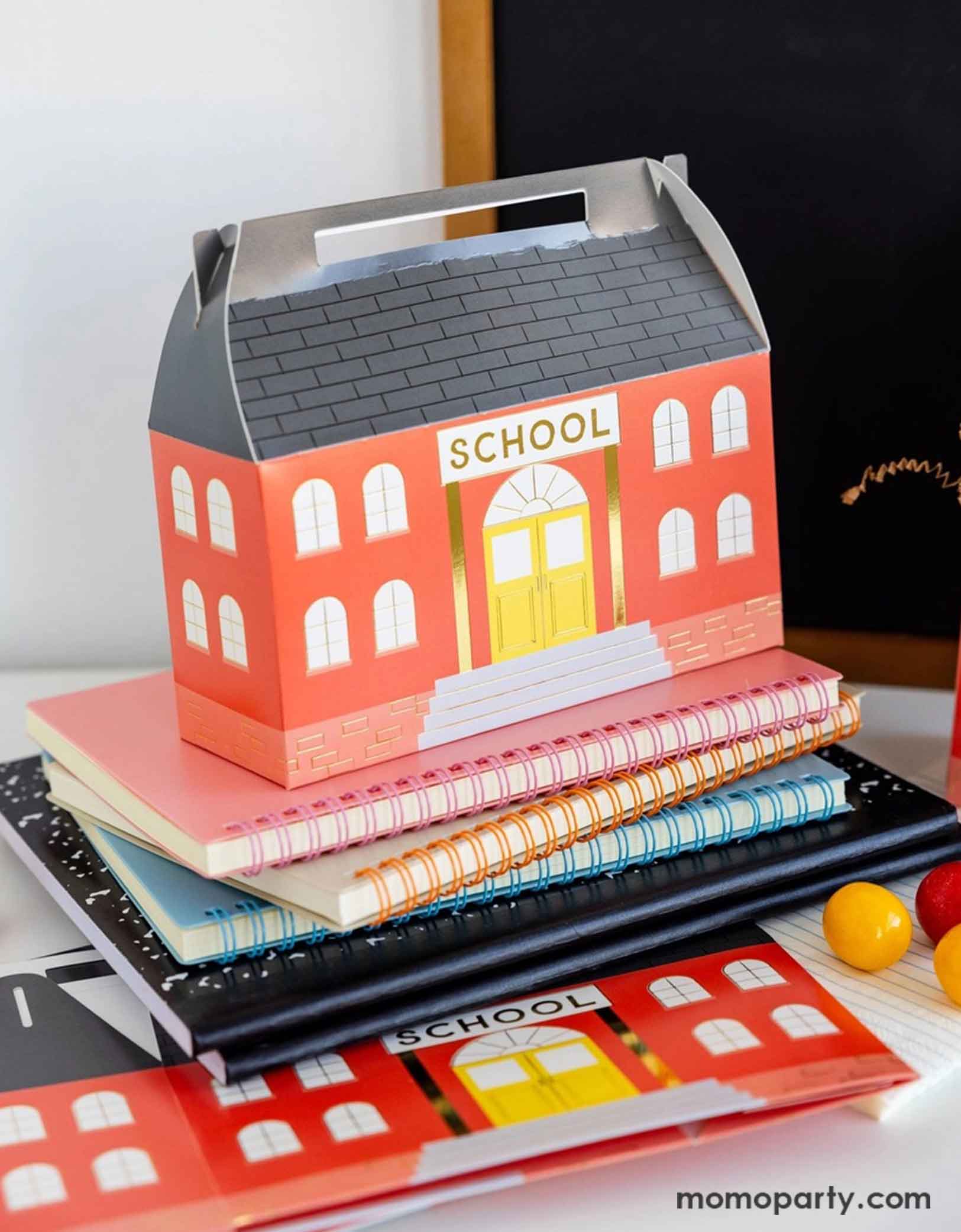 Momo Party's 7" x 2.5" x 5.5" School House Treat Box by My Mind's Eye on a desk with some colorful notebooks and some red and yellow gum balls and in the back there's a small chalkboard, making this an adorable set up for kid's school themed celebration, be it a back to school party or a first day of school celebration.