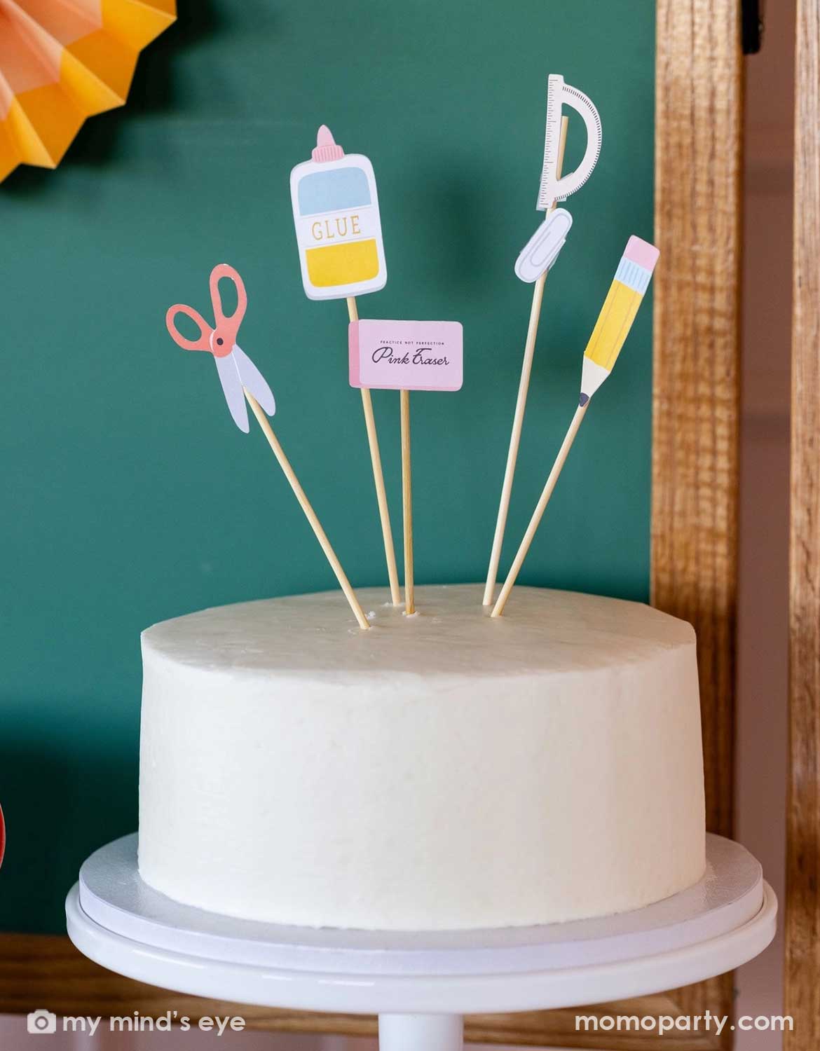 A white cake topped with Momo Party's school-themed cake toppers including scissors, glue, and eraser by My Mind's Eye in front of a school chalkboard.