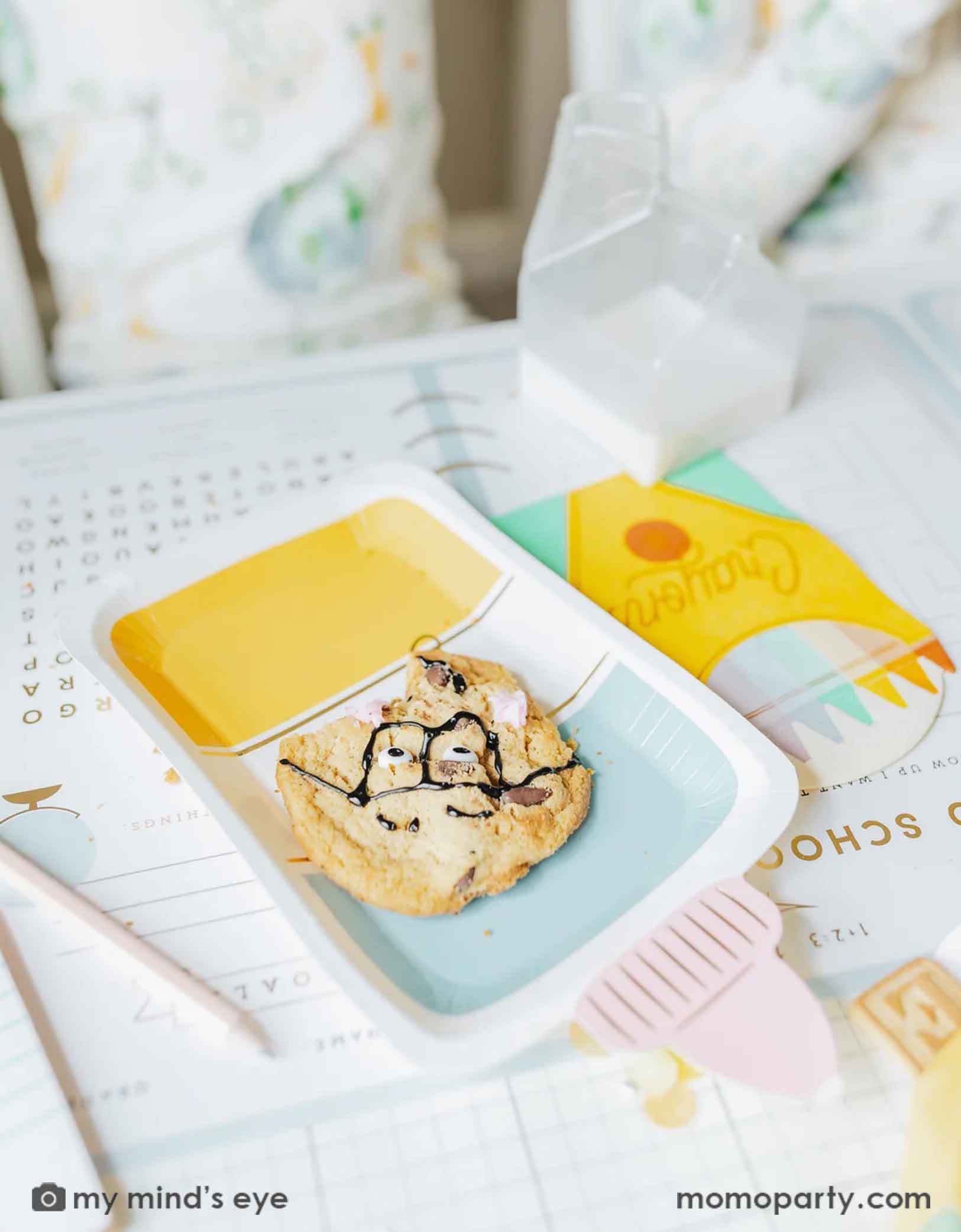 A close up shot of Momo Party's glue bottle shaped party plate by My Mind's Eye with a smart cookie on it. Around the glue bottle plate are some other school themed party supplies including the crayon box shaped napkins, on the back to school activity game placemat, ready for kid's first day of school breakfast celebration!