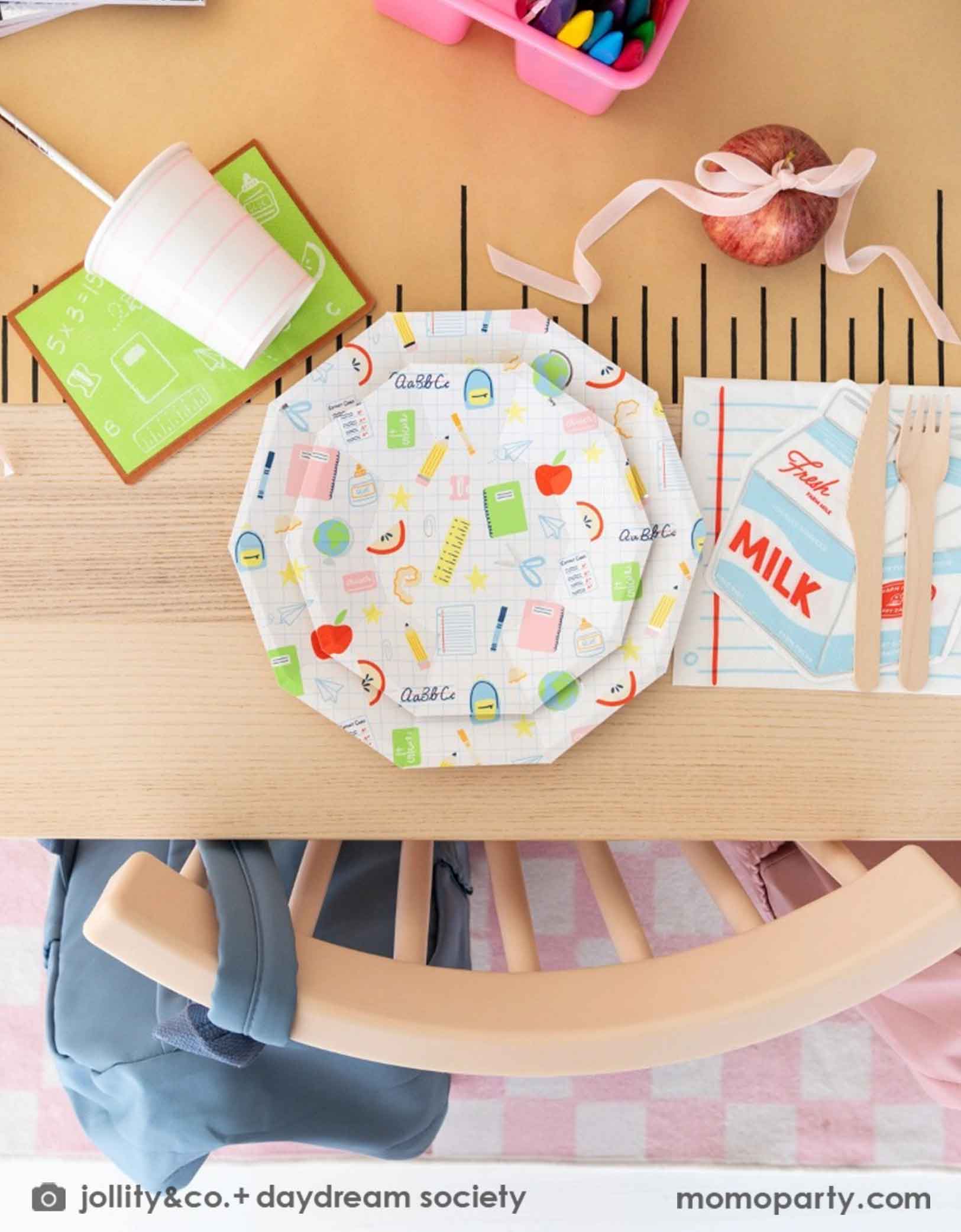 An overhead shot of an adorable kid's school themed party table featuring school supplies themed tablewares from Momo Party including the Smarty Pants milk box shaped napkins, notebook shaped napkins, and pastel pink striped cups, along with school icons patterned plates on a ruler inspired table runner. In the middle of the table there is a pink box with crayons and an apple tied with a pink ribbon, making this an adorable table decor inspiration for kid's first day of school or back to school celebration.