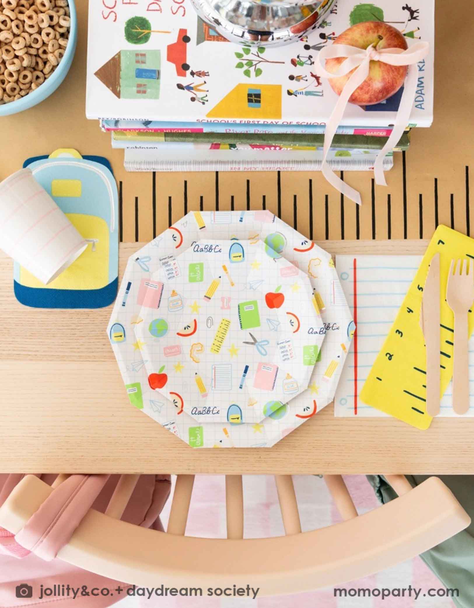 An overhead shot of an adorable kid's school themed party table featuring school supplies themed party tablewares from Momo Party including the Smarty Pants ruler shaped napkins, notebook shaped napkins, and backpack shaped napkins, along with school icons patterned plates, all on a ruler inspired table runner. In the middle of the table there is a pile of books, a bowl of cereal and apples as centerpiece, making this a great decoration inspiration for kid's first day of school or a back to school party.