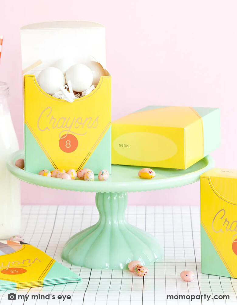 A back to school party table set up in a pink room featuring school themed party supplies from Momo Party including crayon box shaped napkins, crayon box treat boxes by My Mind's Eye on a mint cake stand on a grid table runner next to a milk bottle, with jelly beans around, this makes a great inspiration for a first day of school celebration or kid's back to school parties.