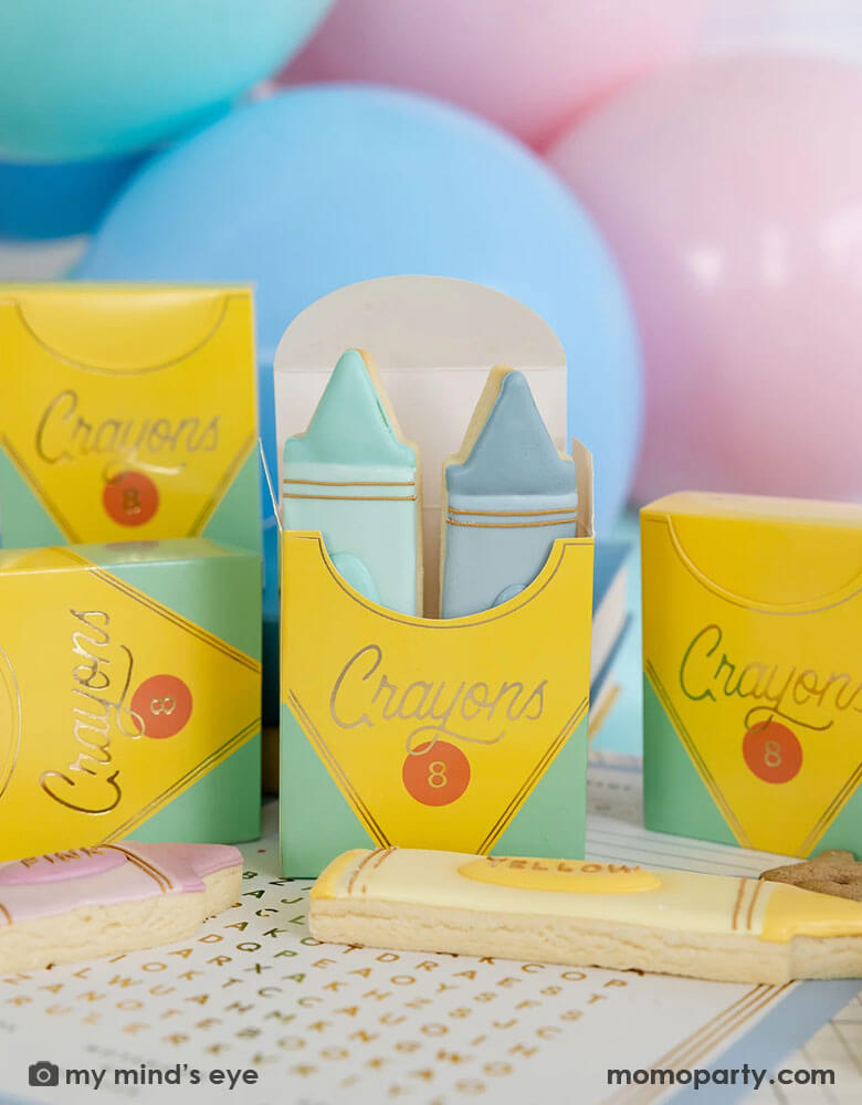 A tableshot of a back to school party featuring crayon shaped treat boxes from Momo Party. The treat boxes are filled with crayon shaped sugar cookies in adorable pastel colors, in the back there are some pastel colored party balloons, making this an adorable idea for kid's first day of school celebration party.
