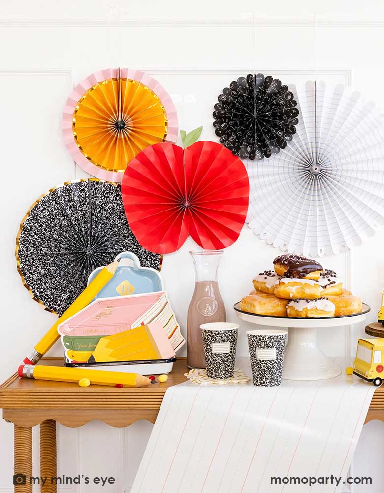 A party table featuring school themed party supplies from Momo Party including stack of books plates, backpack plates, pencil shaped napkins, composition notebook party cups, school bus treat boxes and writing paper table runner around some donuts and milk bottles. The back wall is adorned with back to school party fans by My Mind's Eye including an apple, an pencil, a notebook, an equation pattern and a composition notebook fan. A perfect inspo for kid's back to school, first day of school celebration.