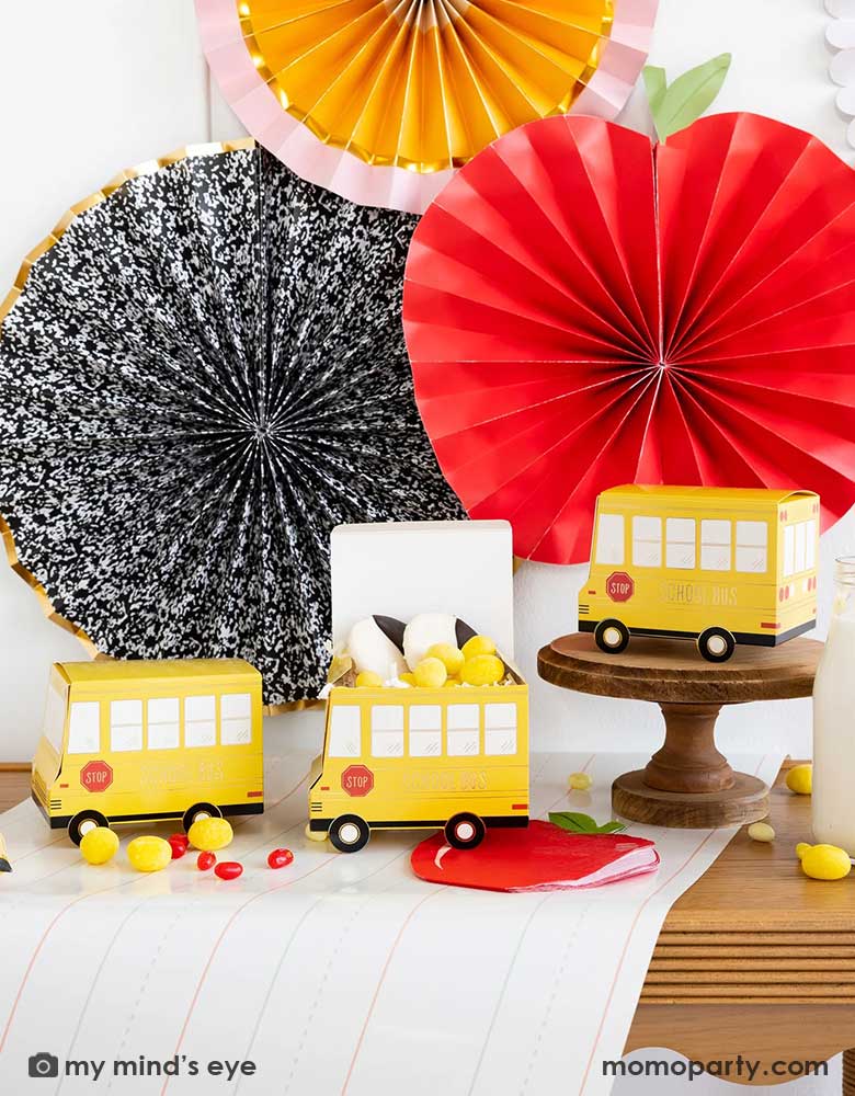 A festive back to school party table features Momo Party's school themed party supplies including school bus shaped treat boxes, apple shaped napkins, writing paper table runner by My Mind's Eye. On the table are some red jelly beans, yellow candies and milk bottles in coordinating colors. In the back there's the back to school party fan hung on the wall, making this a perfect inspiration for kid's back to school party or a first day of school celebration breakfast.
