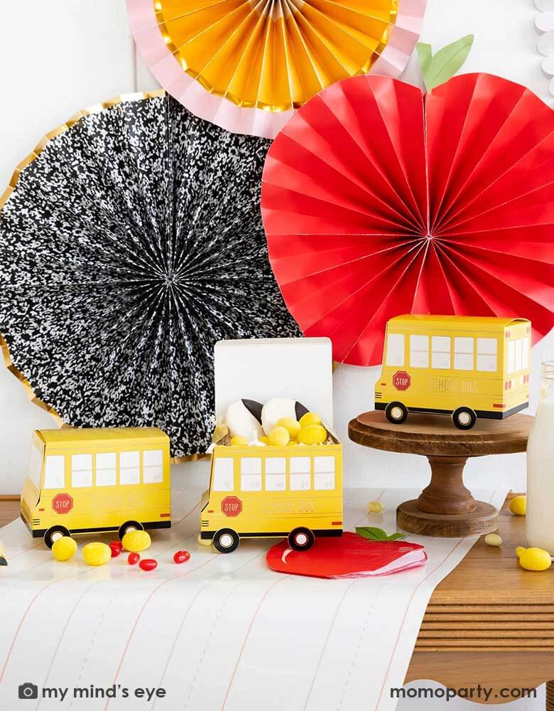 A festive back to school party table features Momo Party's school themed party supplies including school bus shaped treat boxes, apple shaped napkins, writing paper table runner by My Mind's Eye. On the table are some red jelly beans, yellow candies and milk bottles in coordinating colors. In the back there's the back to school party fan hung on the wall, making this a perfect inspiration for kid's back to school party or a first day of school celebration breakfast.