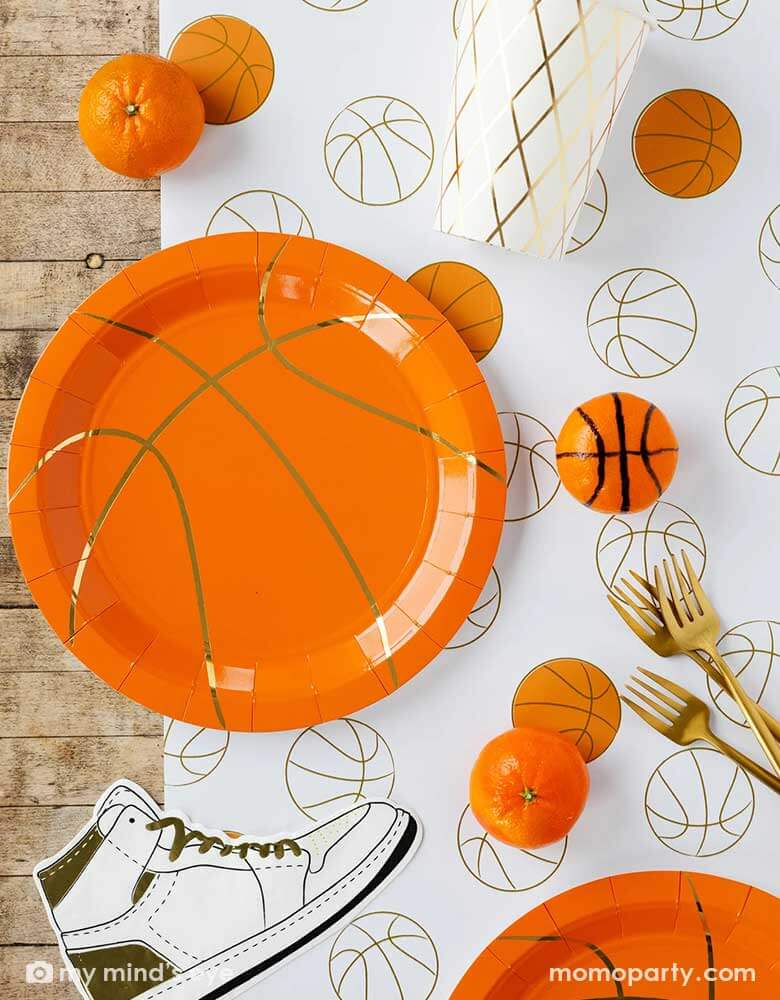 A basketball themed party table features Momo Party's basketball tableware by My Mind's Eye including basketball shaped plates, hightop sneakers shaped napkins, basketball net gold foil party cups and paper basketball table runner, with oranges as snacks on the table - a perfect table setting inspo for kid's basketball themed birthday parties or a fun NBA viewing party!