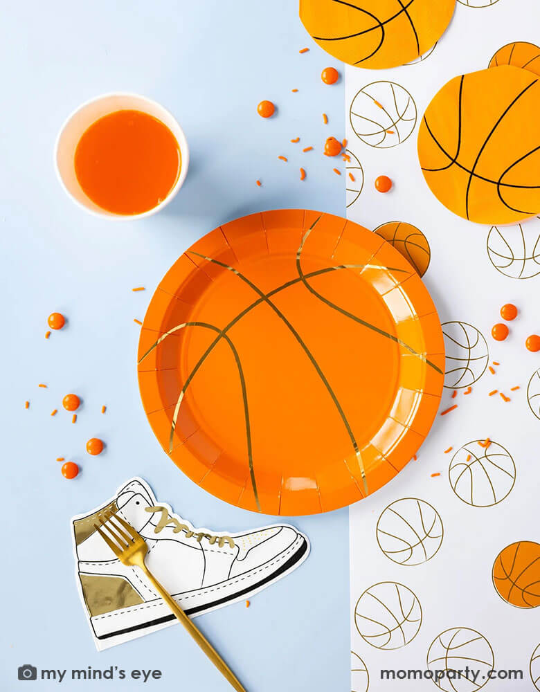 A basketball themed party table features Momo Party's basketball tableware by My Mind's Eye including basketball shaped plates, hightop sneakers shaped napkins, basketball net gold foil party cups and paper basketball table runner, with oranges as snacks on the table - a perfect table setting inspo for kid's basketball themed birthday parties or a fun NBA viewing party!