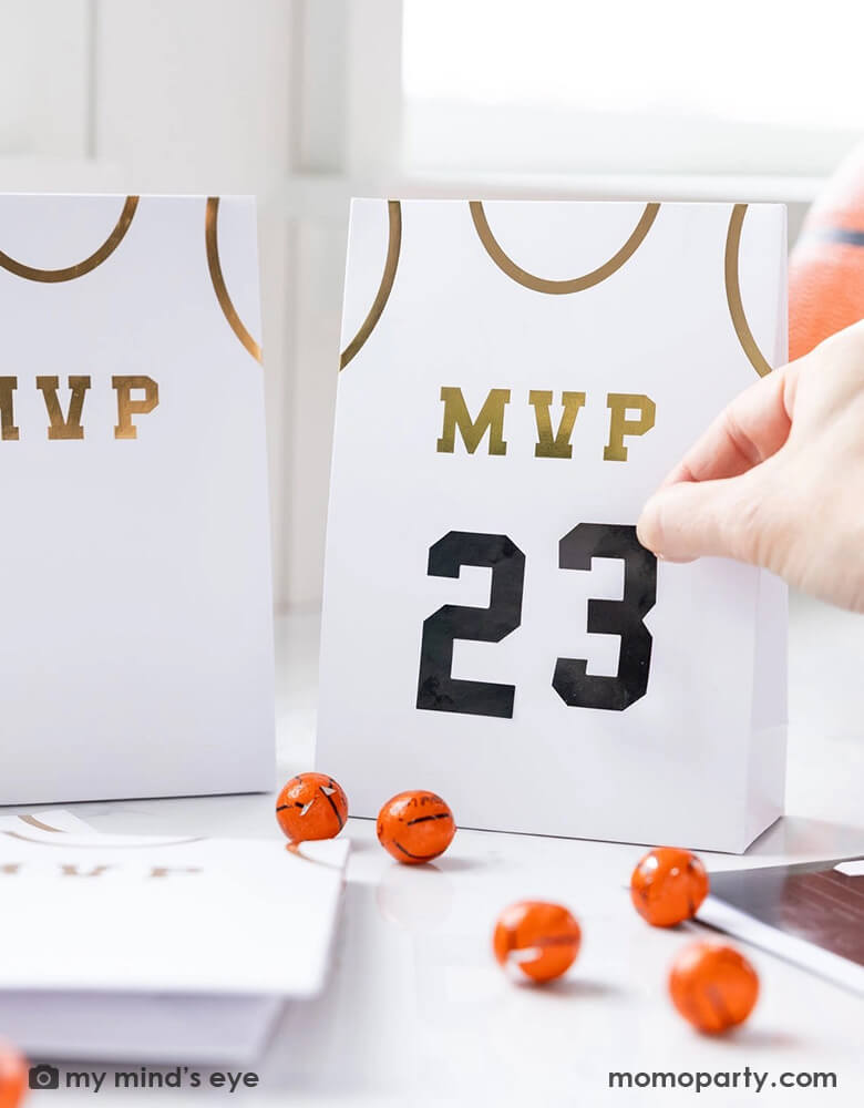A basketball themed party table features Momo Party's basketball treat bags by My Mind's Eye. Comes in a set of 8 treat bags, these basketball jersey shaped treat bags with MVP on them in gold foil are personalizable with the black number stickers. On the table there are some basketball chocolate balls, makes this a perfect idea for party favors at kid's basketball themed birthday party.