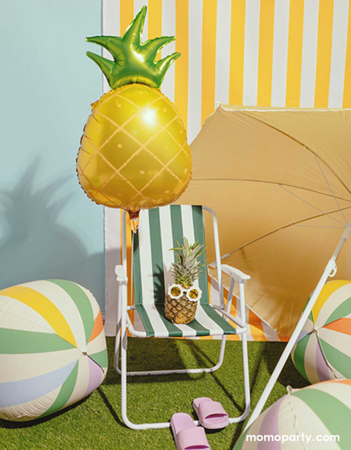 A summer themed party setup with a green striped beach chair, a yellow sun umbrella, pineapple shaped balloon and Momo Party's 19" colorful striped orbz beach ball shaped balloons against a yellow striped wall.