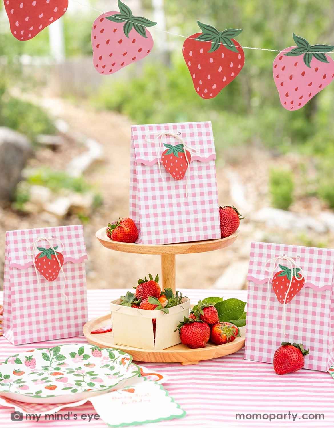 An adorable strawberry-themed party table with strawberry themed decorations and tableware from Momo Party, including pink gingham favor bags with strawberry cutouts, a strawberry party garland with pink and red strawberries, 9" strawberry and vine paper plates and dinner napkins stacked, and strawberries arranged on a wooden cake stand in an outdoor garden setting - a perfect idea for girls "Berry First" strawberry themed first birthday celebration or any strawberry themed parties.