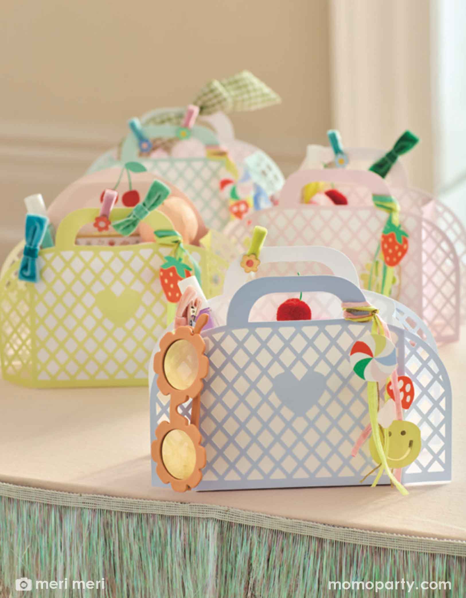 Momo Party's four pastel-colored paper tote baskets filled with party favors and goodies in pink, mint, blue and yellow with decorative keychain charm elements by Meri Meri on a party table adorned with metallic fringe banner.