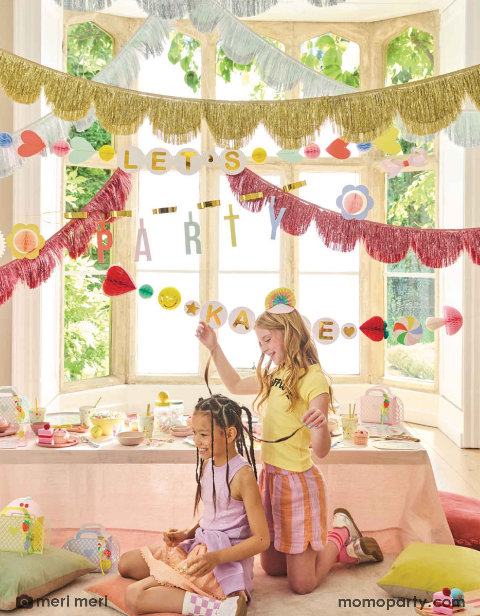 Two pre-teen girls in a room decorated with colorful Momo Party's Charm Party Collection by Meri Meri including festive banners, metallic fringe garlands, and a table of charm party themed tablewares including plates, napkins and cups. A perfect set up for girl's sleepover party or a BFF gathering.