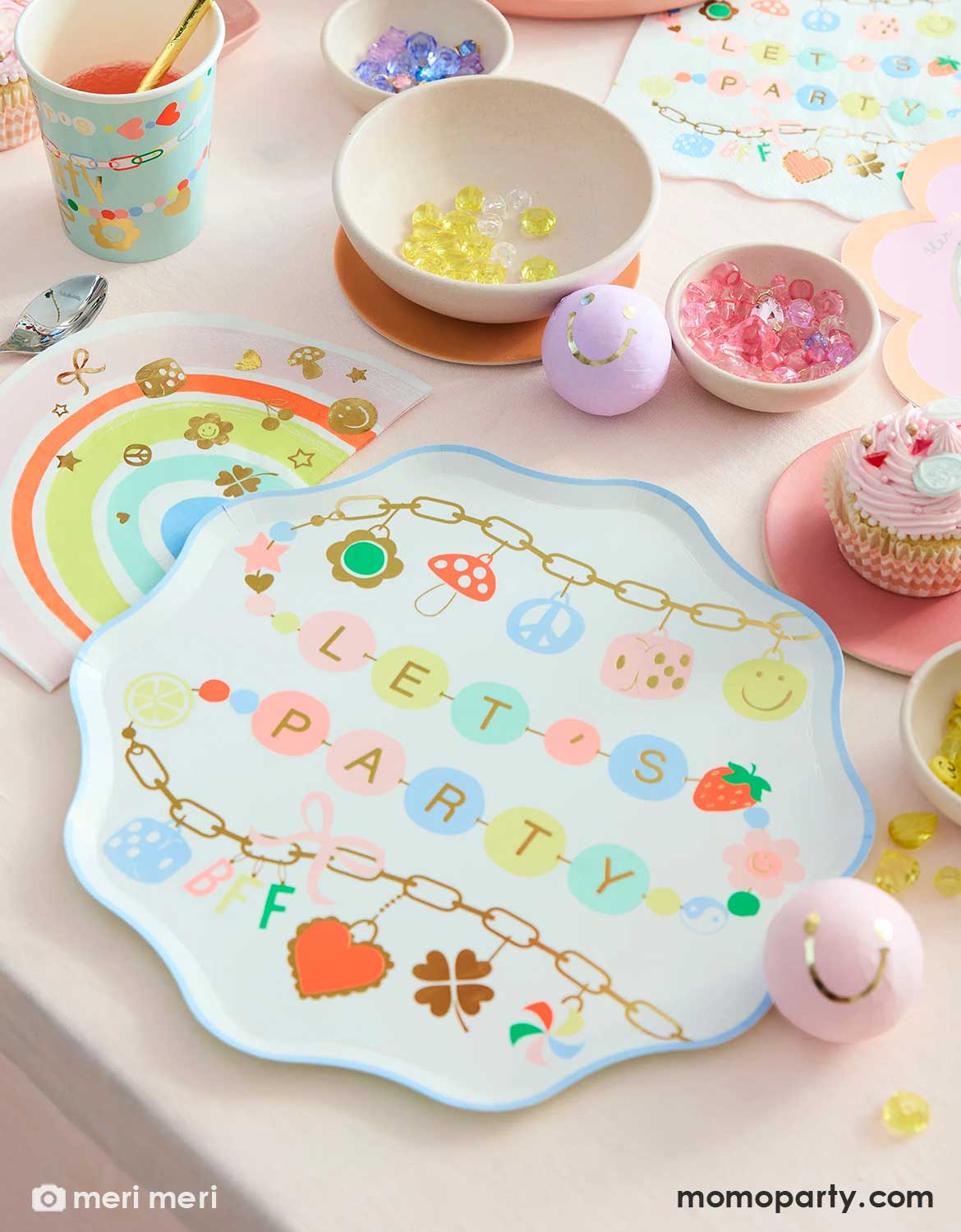 An overhead shot of a girly charms themed birthday party table set up with Momo Party's charm party tablewares including dinner plates, napkins, party cups and rainbow shaped napkins in pastel colors. These tablewares are surrounded by colorful treats, art and crafts supplies, beads and party favors, making this a fun set of for girl's sleepover parties, besties birthdays, Taylor Swift or pop star themed celebrations and more.