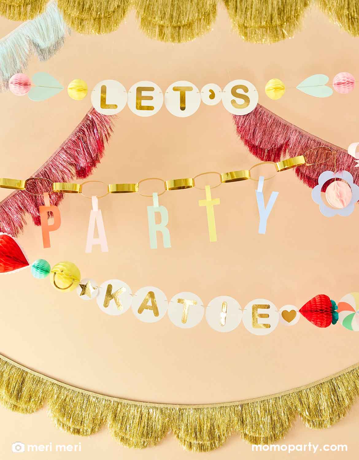 Momo Party's colorful Charm Party Garland Set by Meri Meri with 'Let's Party' message on the banner with decorative elements including heart shaped honeycombs, bows, daisies and a personalizable pennants for kid's name "Katie" on a beige background. This festive banner set is perfect for kid's sleepover parties or a BFF themed birthday celebration.
