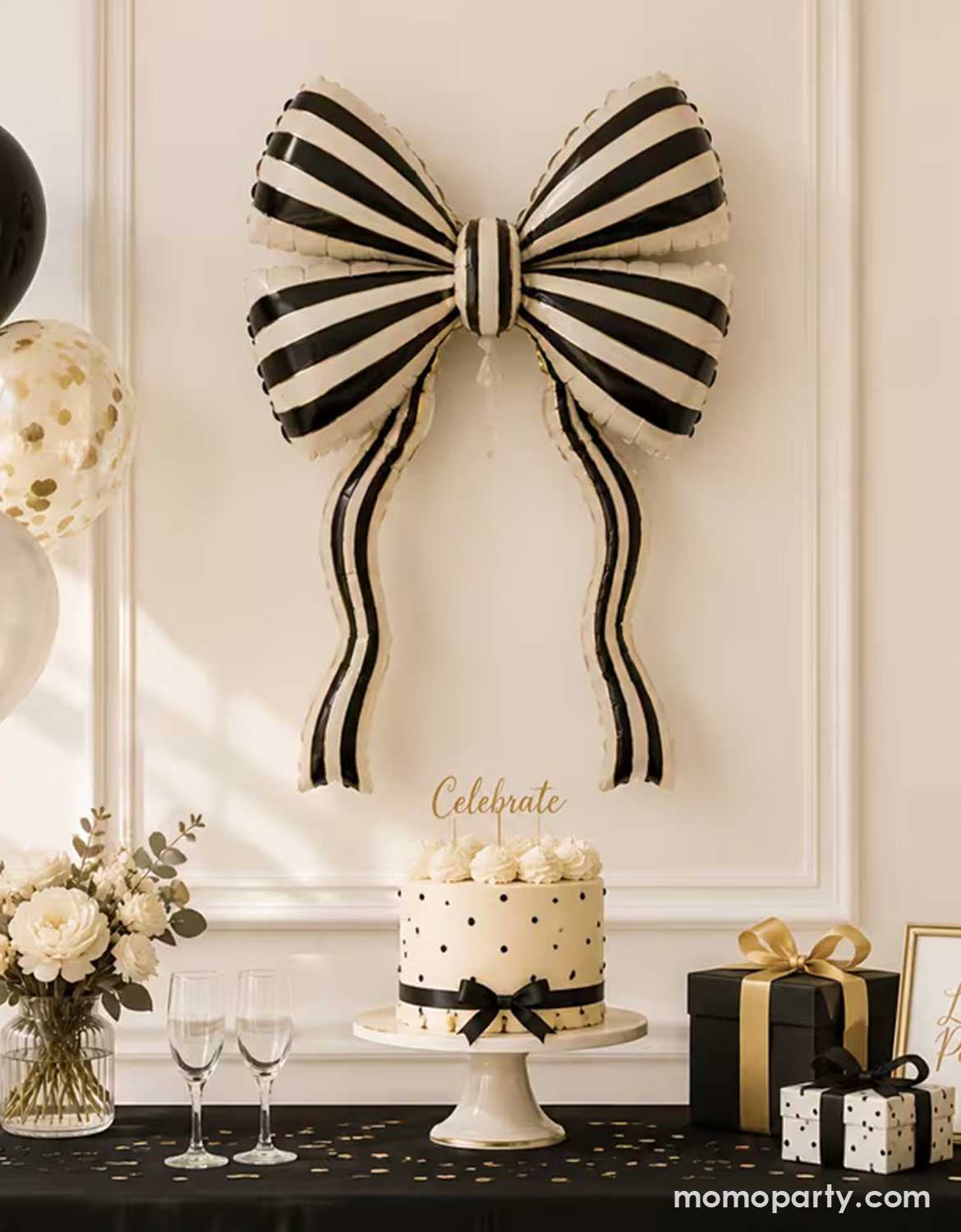 Elegant black and white striped bow shaped foil balloon styled as the centerpiece of a chic coquette bow themed celebration, featuring a black and white bow birthday cake, gold accents, confetti, flowers, champagne glasses, gift boxes with ribbon bows, and coordinating black, white, and gold party decorations by Momo Party.