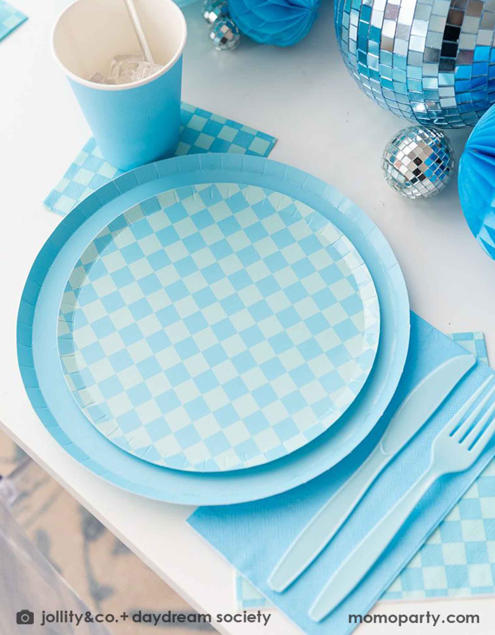 A blue themed party table features Momo Party's Check it! Blue and Blue Checkered Tableware Collection by Jollity & Co. including 10" blue dinner plates, 8" blue checkered dessert plates, 6.5" large napkins, and 5" blue checkered small napkins, blue plastic utensils and solid blue party cups, with blue honeycomb and disco ball decorations as the centerpiece for the party table.