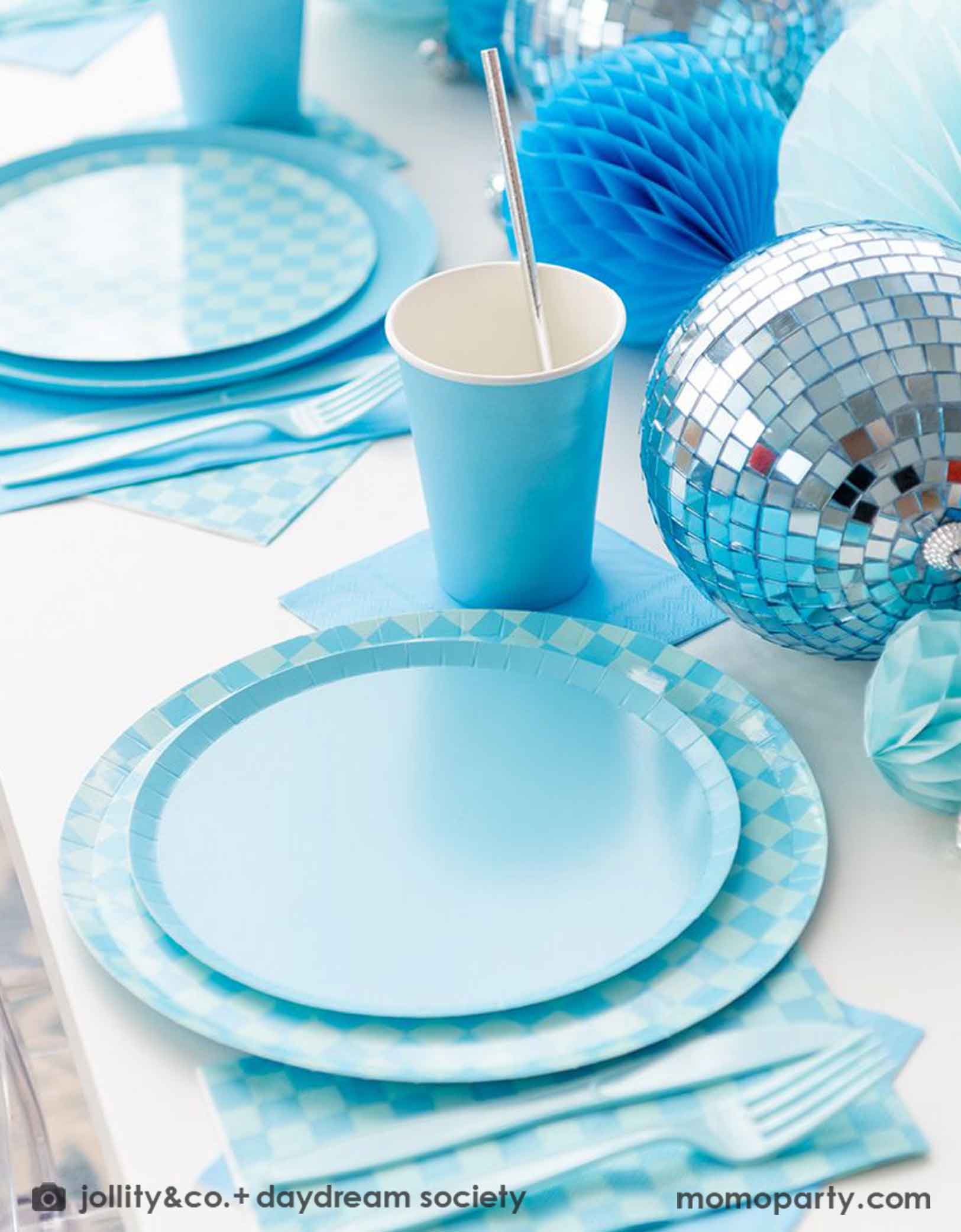 A blue themed party table features Momo Party's Blue and Check it! Blue Checkered Tableware Collection by Jollity & Co. including 10" blue checkered dinner plates, 8" blue dessert plates, 6.5" blue checkered large napkins, blue plastic utensils and solid blue party cups, with blue honeycomb and disco ball decorations as the centerpiece for the party table.